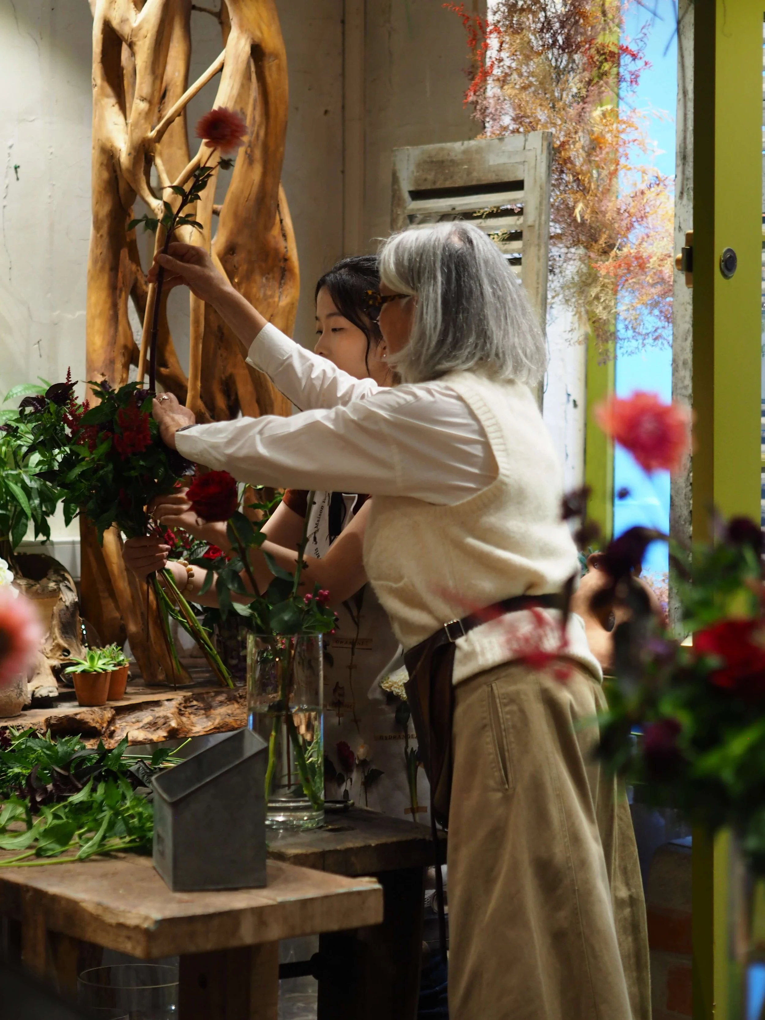 Deux femmes, une avec des cheveux gris et une plus jeune, préparent ou arrangent des fleurs dans un atelier floral. Elles sont concentrées sur leur activité, entourées de fleurs, de vases et de matériaux de décoration.