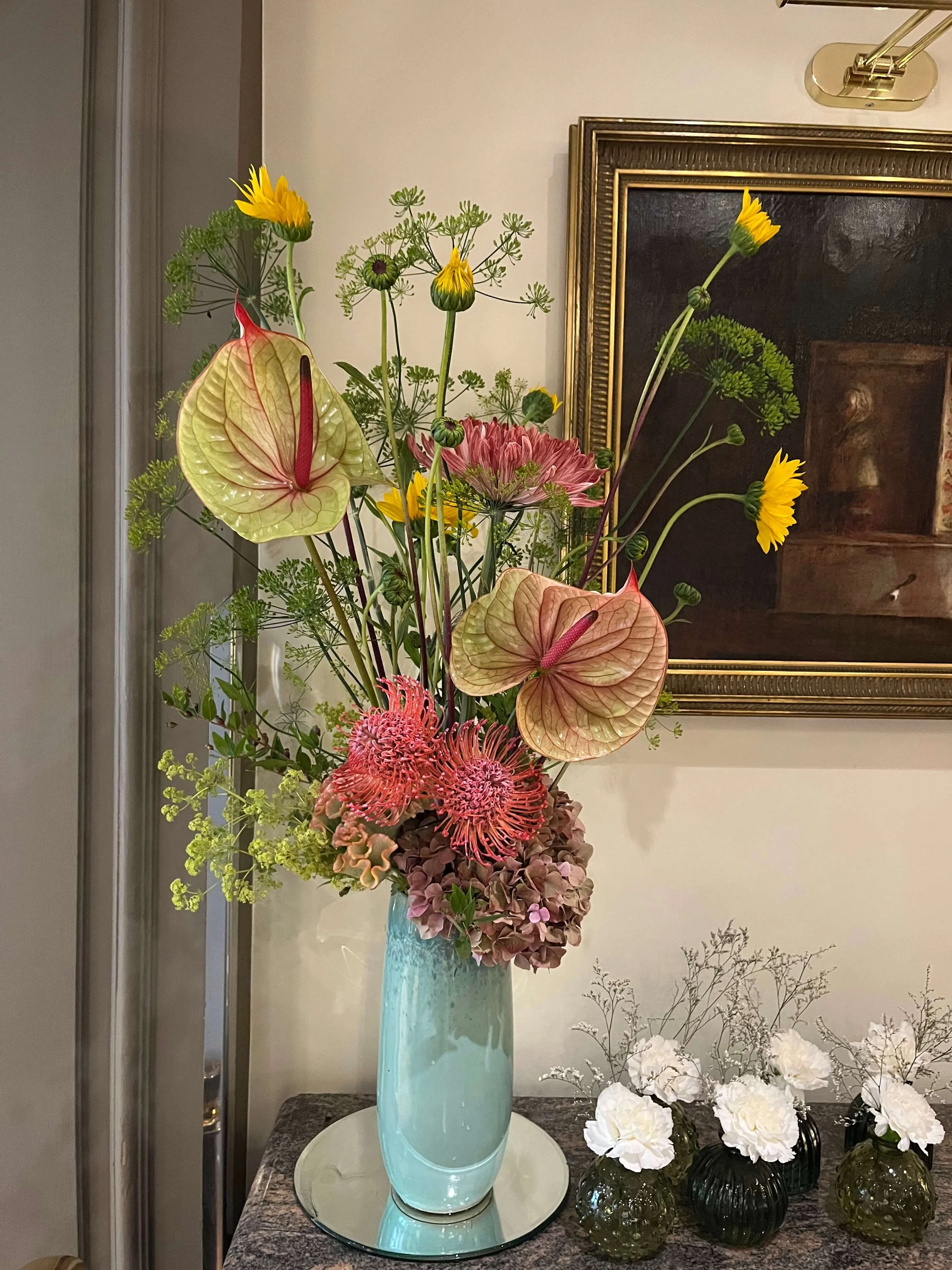 Un arrangement floral avec des fleurs colorées dans un vase turquoise, placé sur une table en marbre, avec un tableau encadré en arrière-plan.