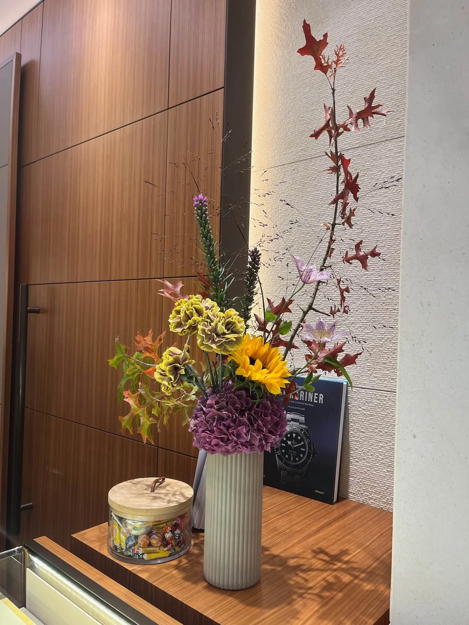 Un vase blanc avec des fleurs colorées, notamment des hortensias mauves, des tournesols, des roses lavandes et des feuilles rouges, est placé sur une surface en bois. À côté, il y a un bocal transparent avec des bonbons. Derrière, un livre sur une mo