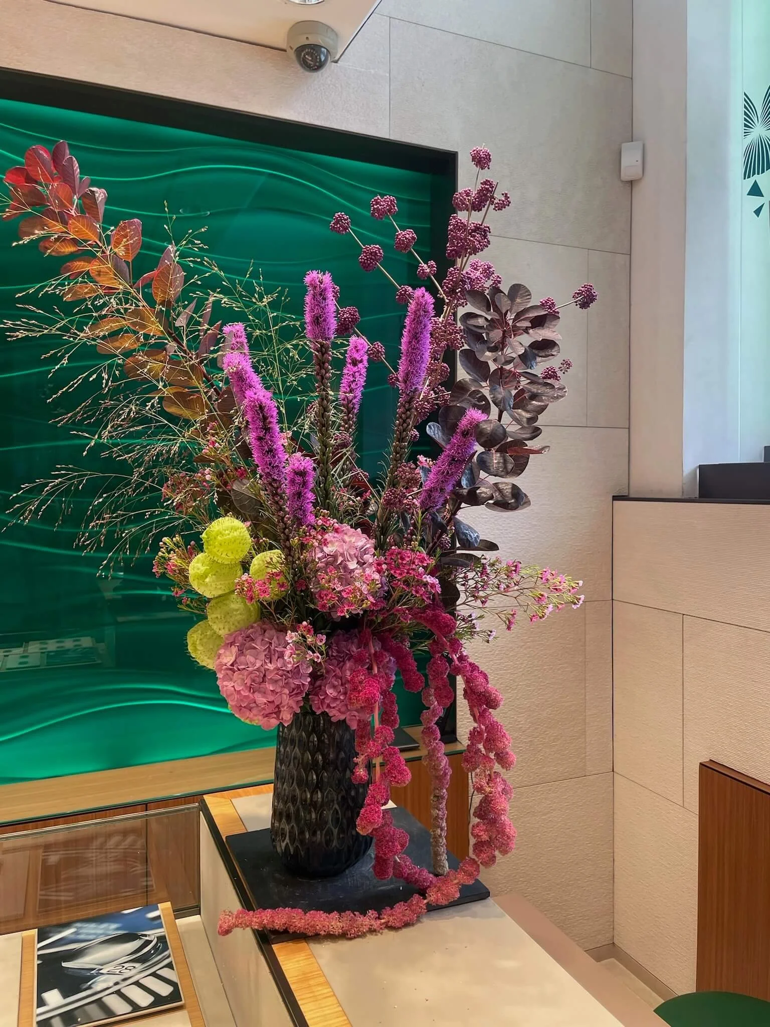 Grand arrangement floral avec des fleurs violettes, roses, vertes et brunes dans un vase noir, placé sur une table beige dans un intérieur moderne.