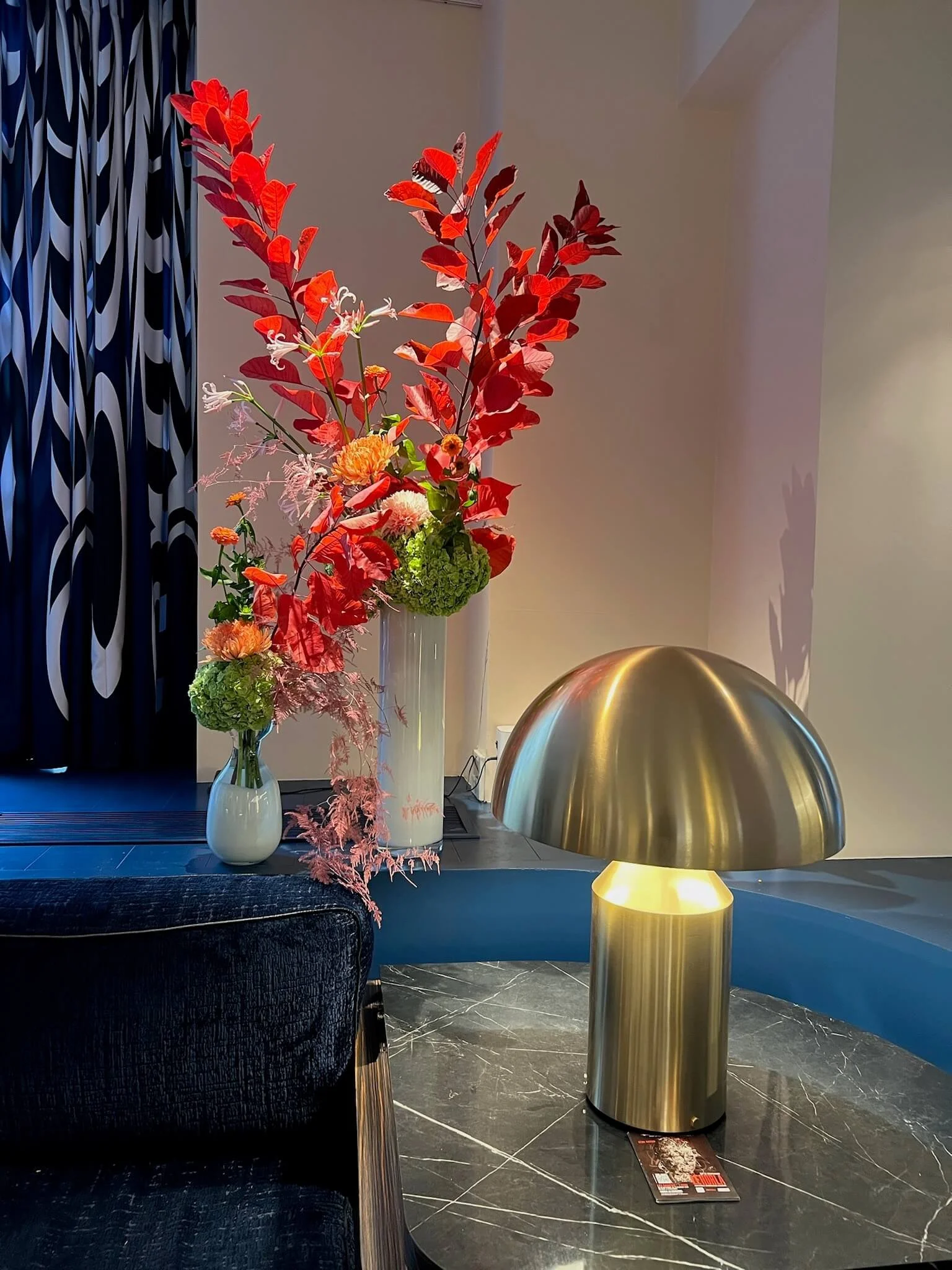 Grand vase rectangulaire avec des fleurs rouges, blanches, jaunes et vertes, avec un lampadaire en forme de dôme doré sur une table en marbre noir.