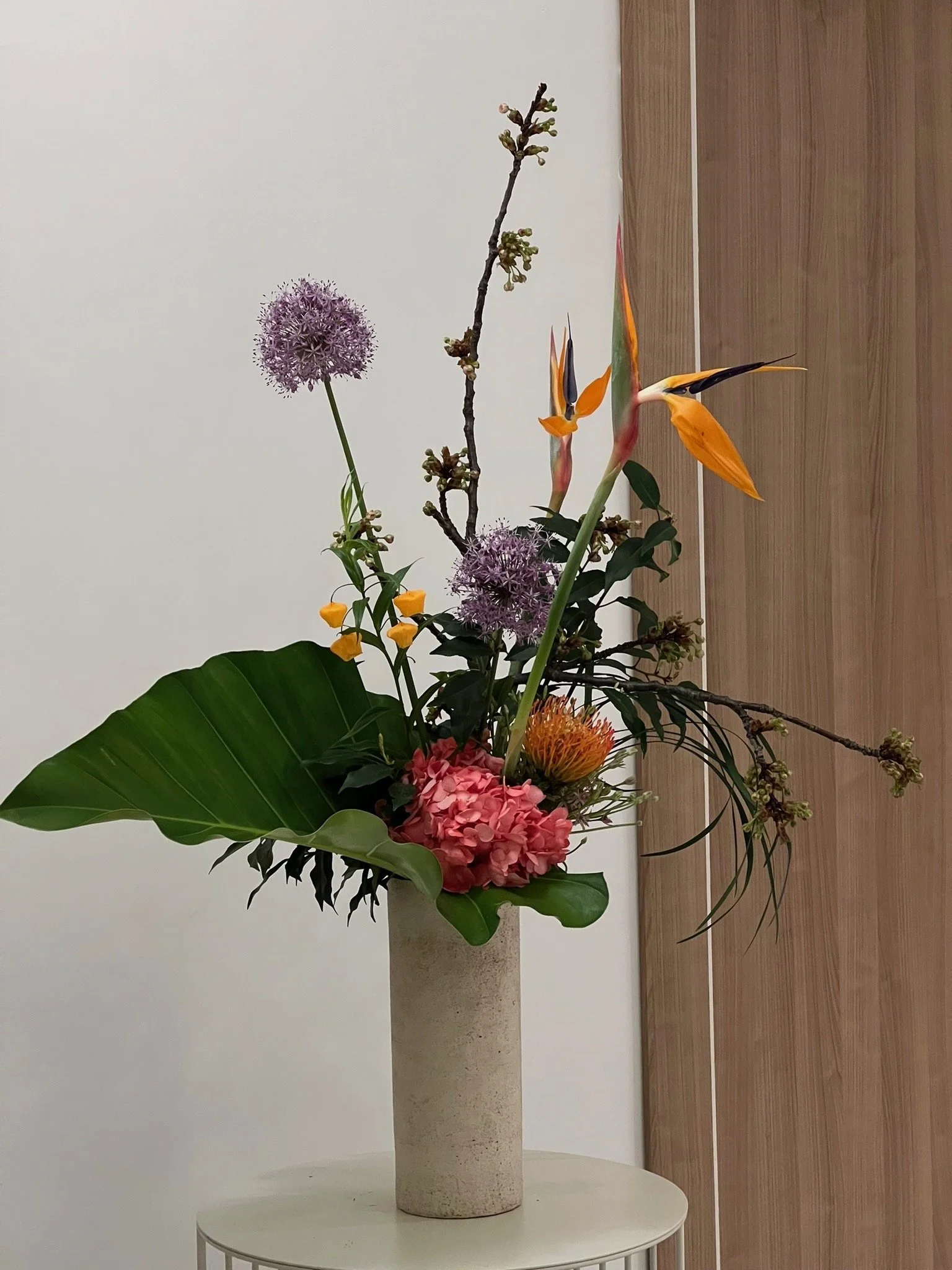 Composition florale avec des orchidées orange, des hydrangeas roses, des alliums violets, des branches et des feuilles vertes dans un vase en céramique dans une pièce moderne.