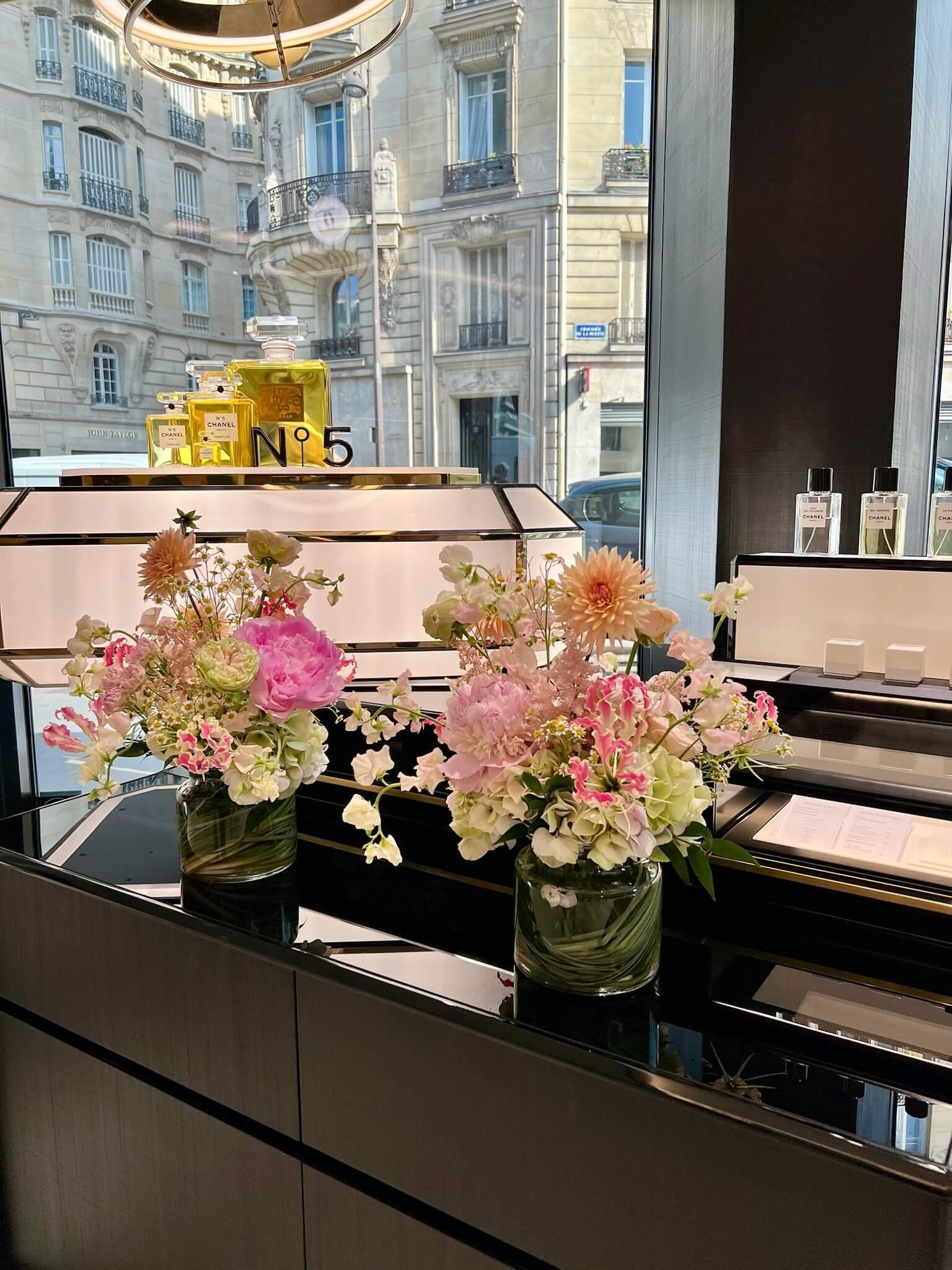 Vase avec des fleurs pink, blanc et vertes sur un meuble noir dans une boutique de parfum avec des bouteilles de parfum Chanel. Fenêtres offrant une vue d'un bâtiment haussmannien avec des balcons en fer forgé.