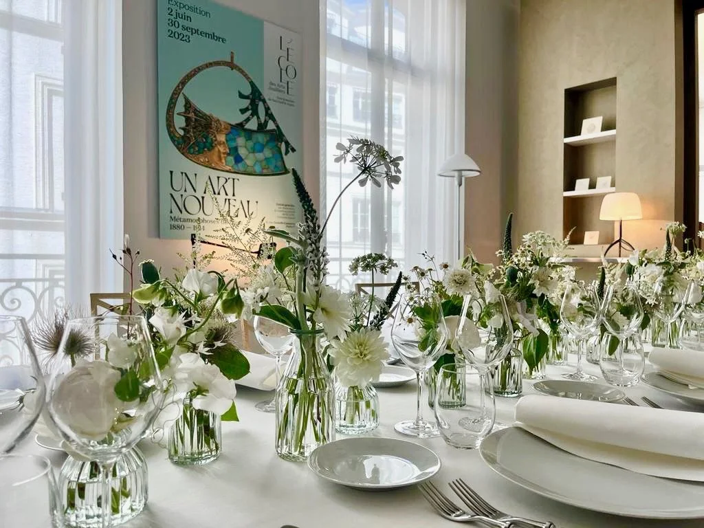 Une table élégante avec des bouquets de fleurs blanches dans des vases en verre, positionnée dans une pièce lumineuse avec une affiche d'exposition sur le mur.
