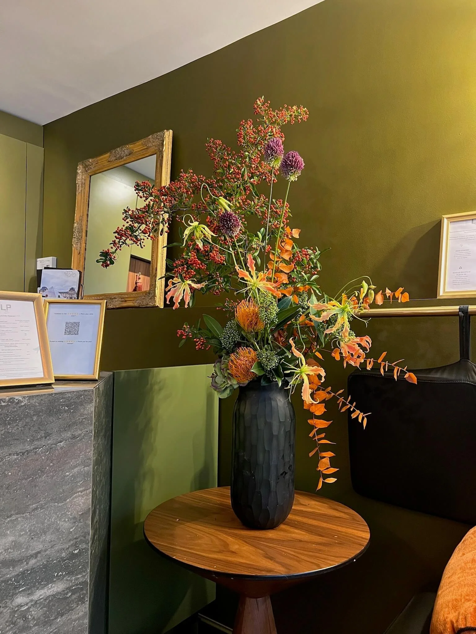 Un vase noir contenant un arrangement floral coloré sur une table en bois dans un intérieur avec mur vert et miroir en bois.