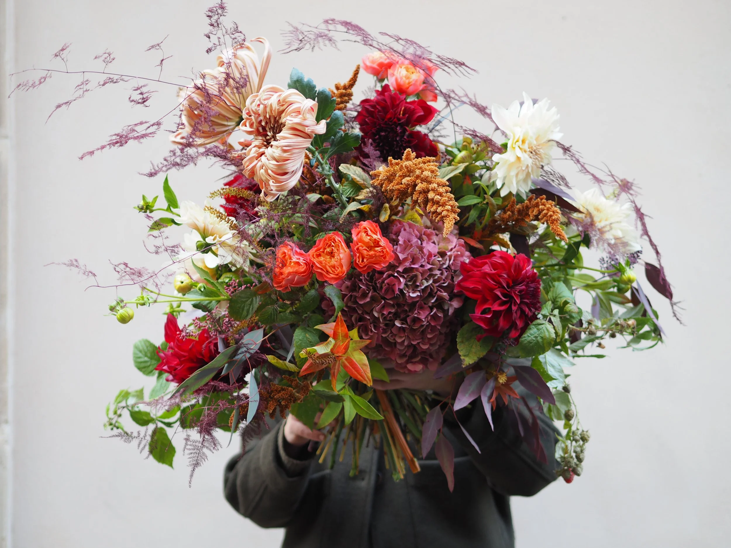 Personne tenant un grand bouquet de fleurs colorées et variées devant leur visage contre un fond neutre.
