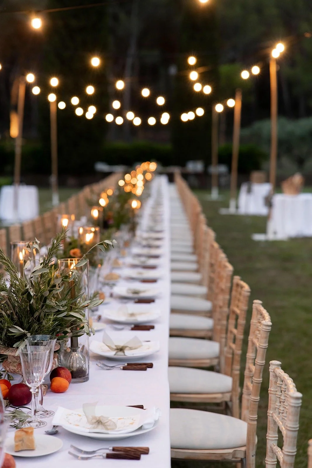 Une grande table dressée pour un repas en extérieur, décorée avec des bougies et des fruits, sous un éclairage suspendu en soirée.