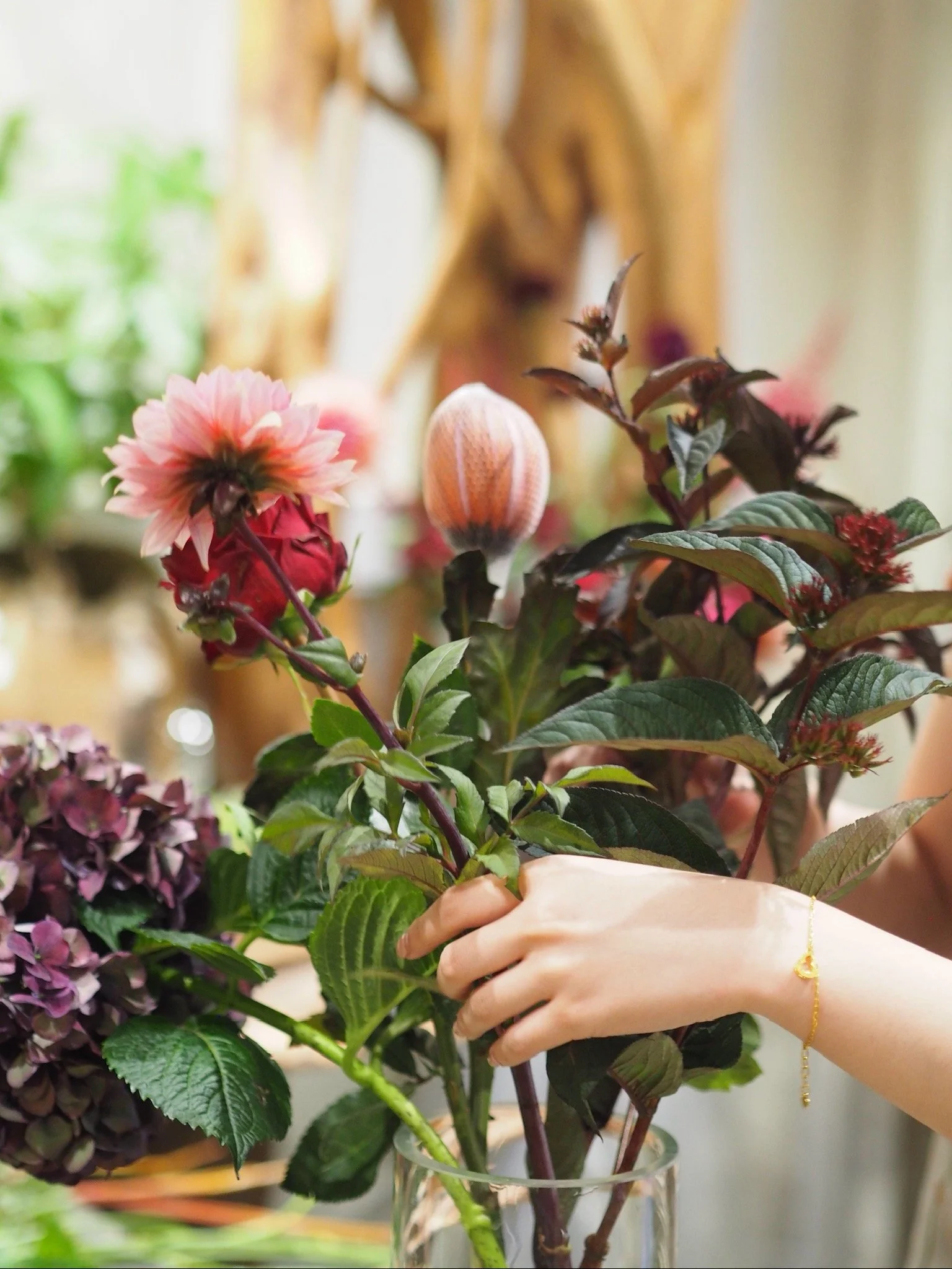 Une main tient un bouquet de fleurs variées, notamment un dahlia rose et une rose rouge, avec un arrière-plan flou de plantes vertes et d'un décor intérieur.