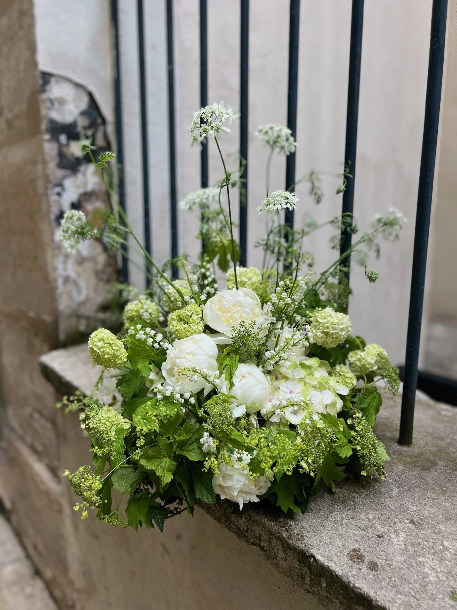 Composition florale blanche avec pivoines, hortensias et petites fleurs sauvages, posée sur un rebord en pierre devant une grille en fer et un mur abîmé.
