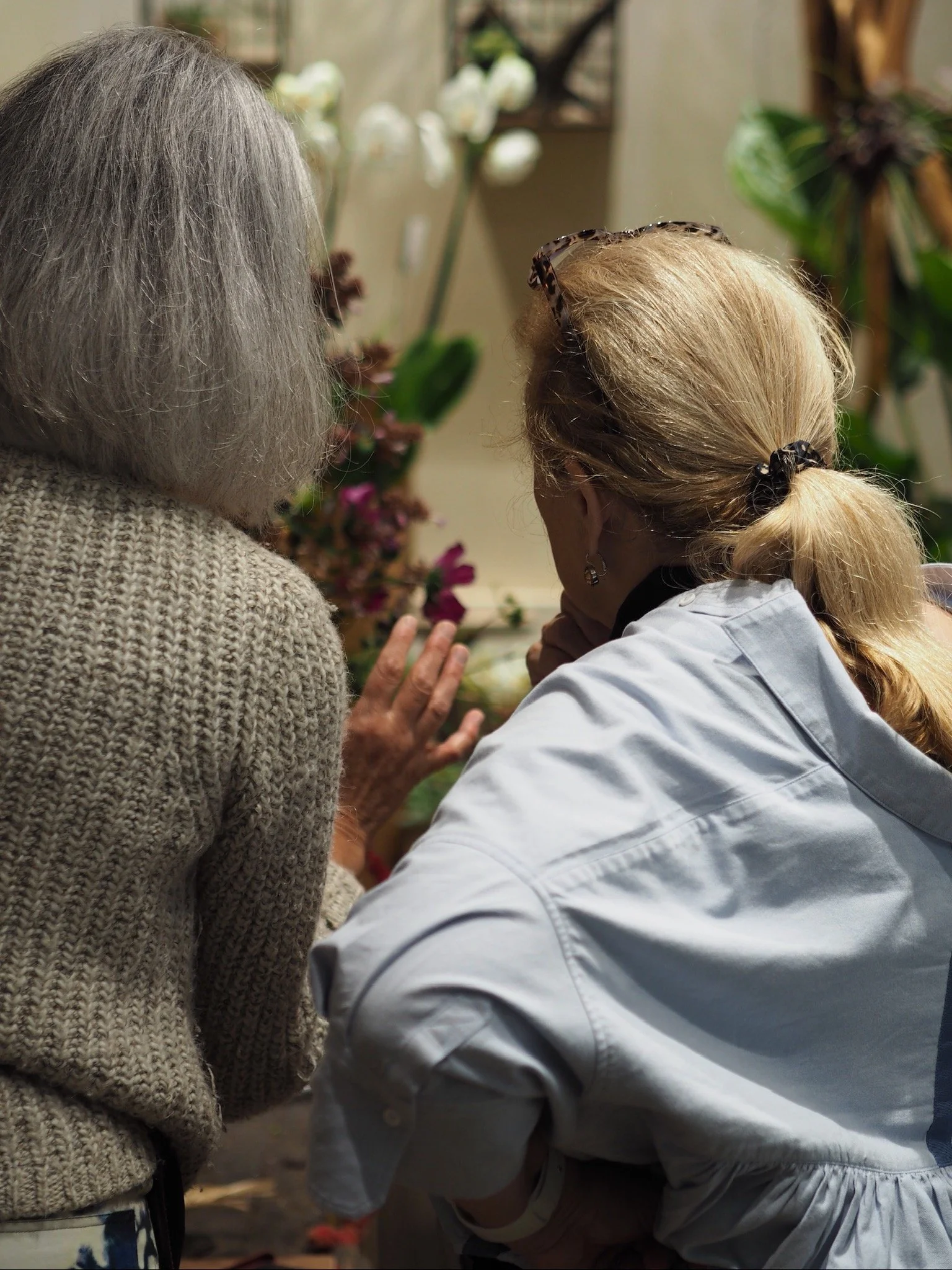 Deux femmes en conversation, l'une porte une veste blanche, l'autre un pull en laine beige, entourées de plantes et de fleurs dans un environnement intérieur.