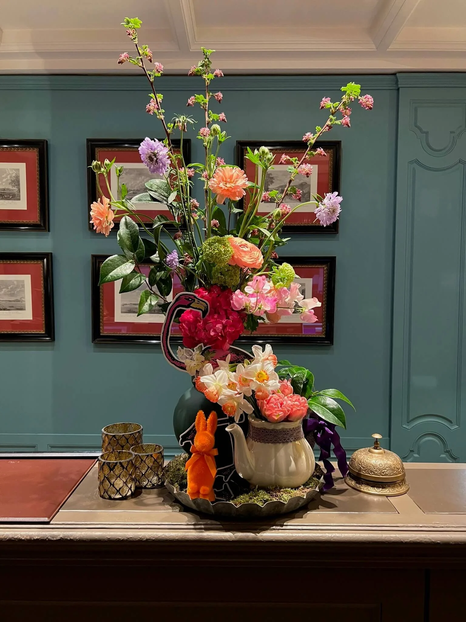 Vase de fleurs colorées avec décoration en forme de flamant rose, lapin orange, et boîte à bonbons en or sur une table en bois.