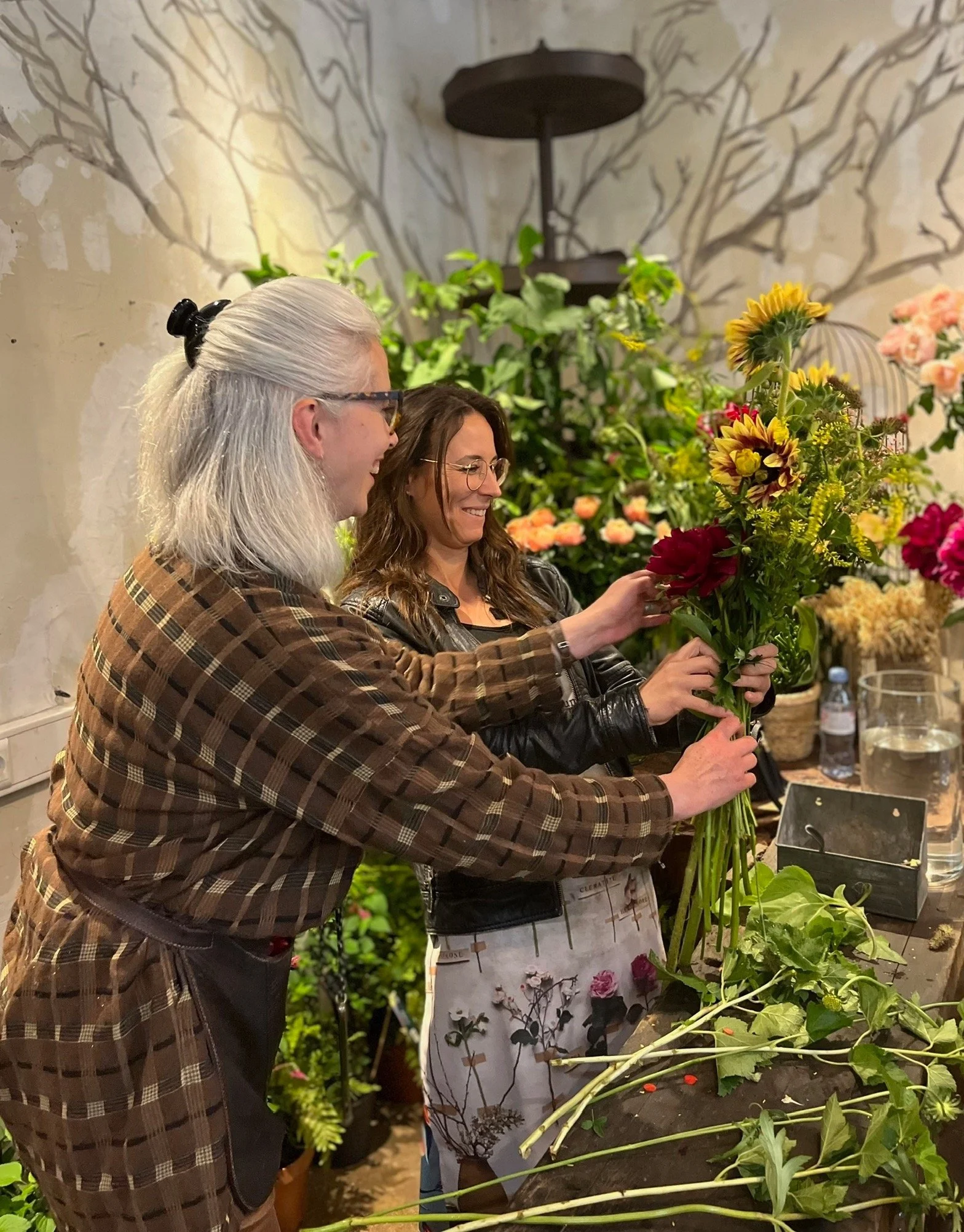 Deux personnes, une avec des cheveux gris longs attachés en queue de cheval et une femme avec des cheveux bruns, sourient et manipulent des fleurs dans un atelier de fleurs, entourées de diverses fleurs colorées et de végétation.