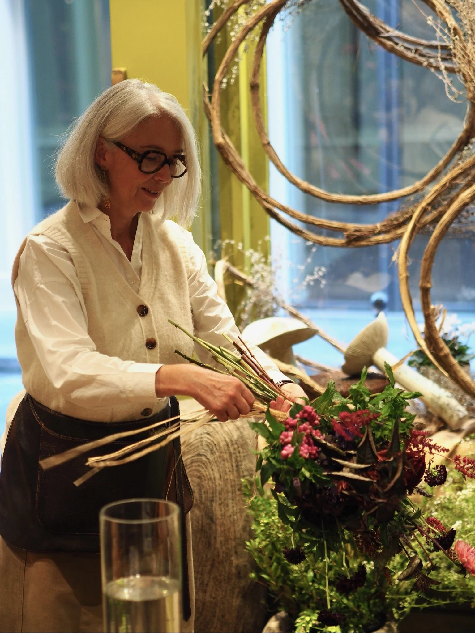 Une femme, portant des lunettes, prépare un arrangement floral dans un intérieur décoré avec des éléments naturels et des champignons en plastique ou en céramique.