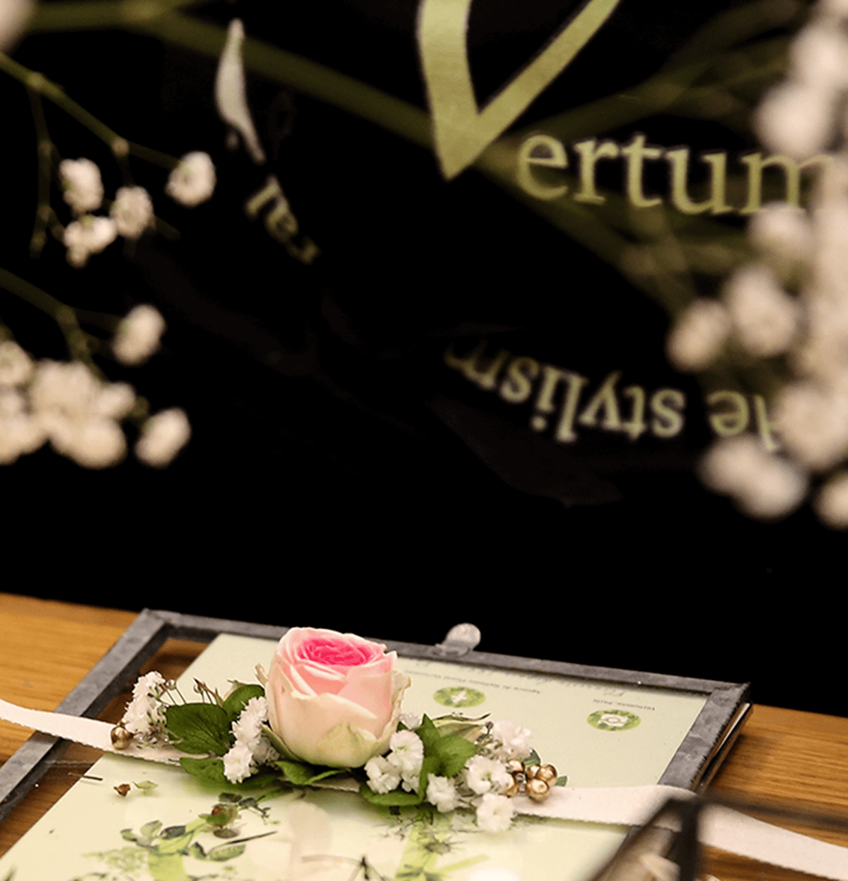 Une petite décoration florale avec une rose rose pâle entourée de petites fleurs blanches et de feuilles vertes, posée sur une carte ou un cadre en bois sur une table en bois.