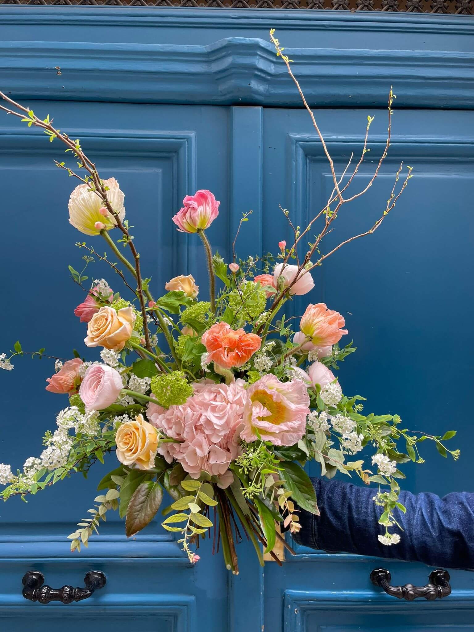 Bouquet de saison aux teintes pastel avec roses abricot, hortensias, pavots et feuillages texturés, tenu devant une porte bleu profond.