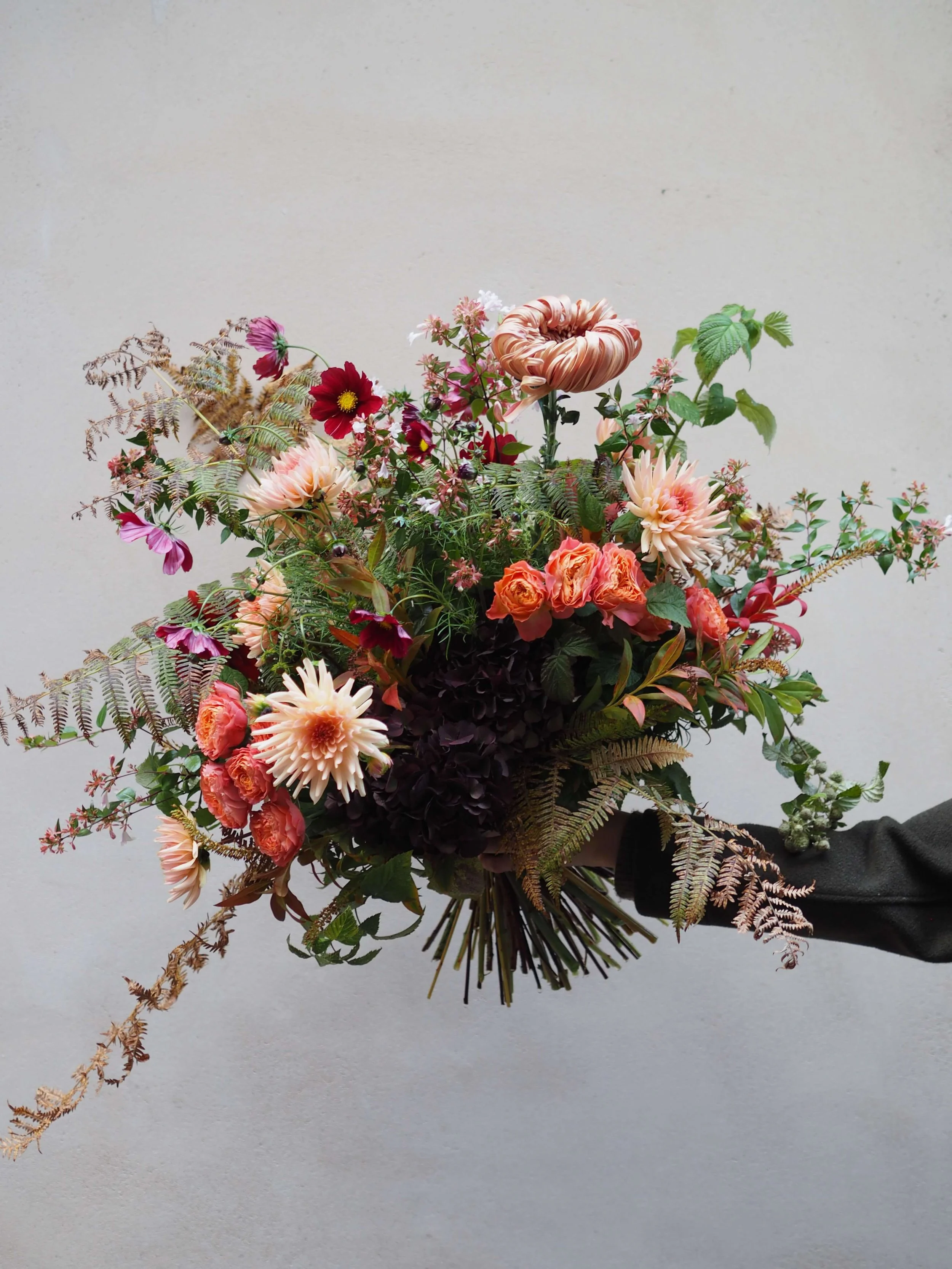 Un bouquet de fleurs variées avec des teintes orange, blanches, rouges et vertes, tenu par une personne portant une veste noire, contre un mur blanc.