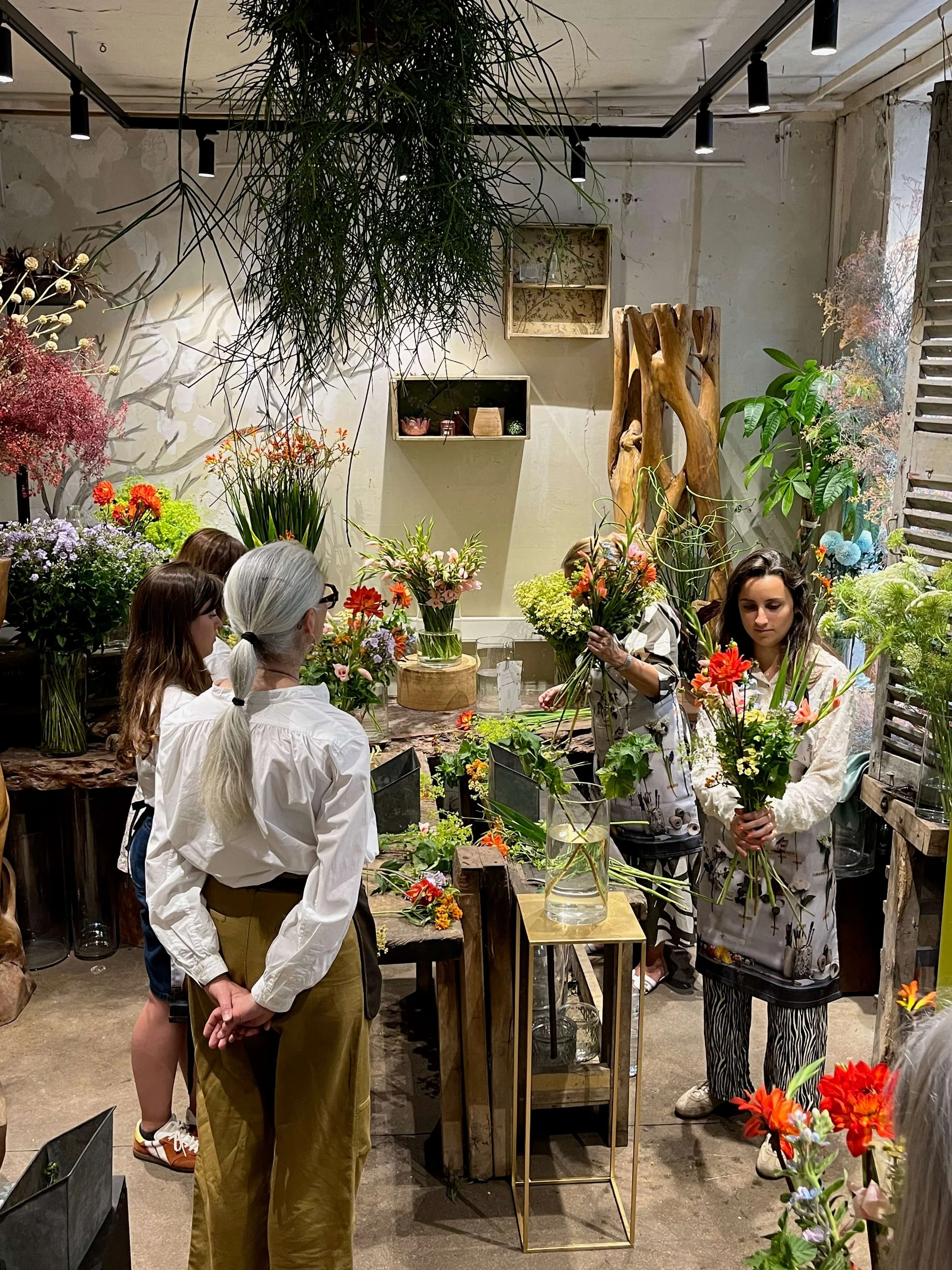 Groupes de personnes dans un atelier de fleurs, manipulant et examinant des bouquets de fleurs colorées, dans un espace décoré avec des plantes et des fleurs.