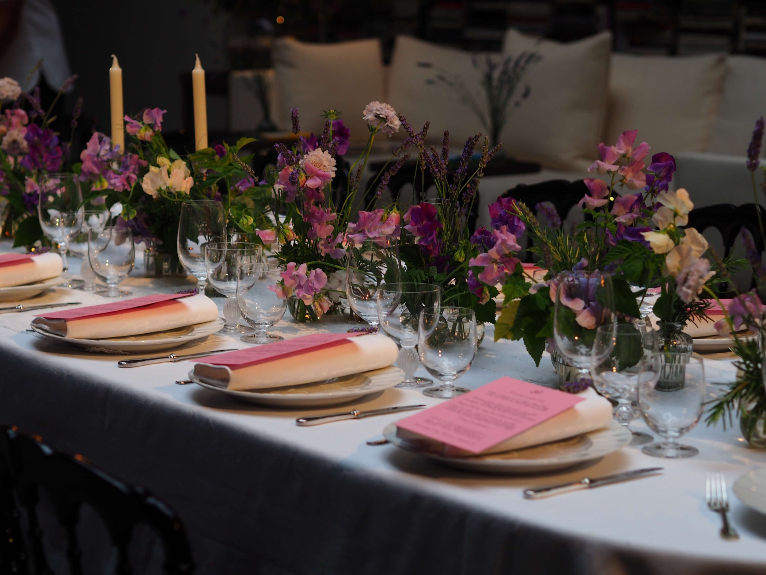 Une table décorée avec un centre de fleurs composé principalement de fleurs roses, violettes et blanches, accompagnée de bougies longues et de vaisselle élégante, dans un cadre intérieur.
