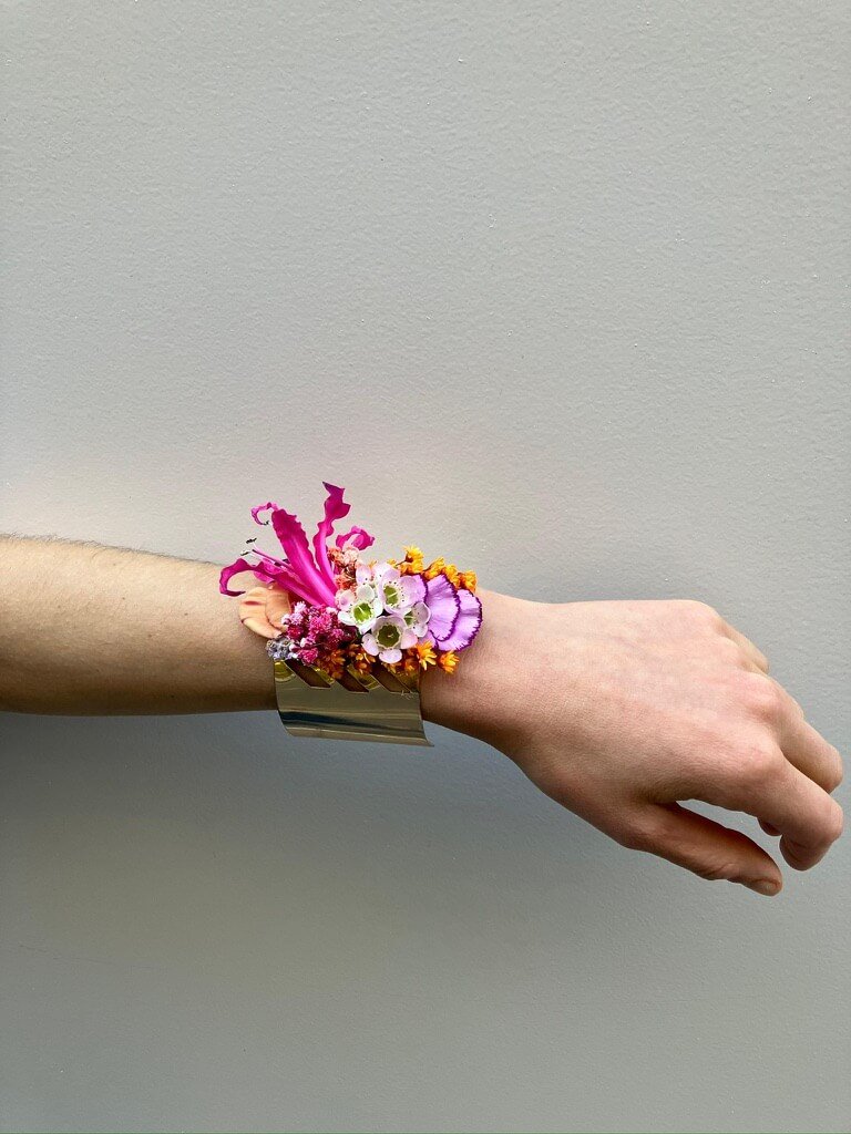 Poignet décoré avec un bracelet floral coloré, comprenant des fleurs roses, violettes, oranger et blanches, porté sur un bras contre un mur gris.