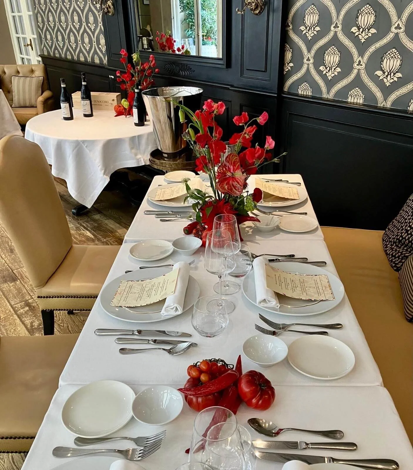 Table de restaurant élégante avec vaisselle, verres à vin, couverts, et une décoration florale rouge au centre, en salle ornée de papier peint à motifs noirs et dorés.