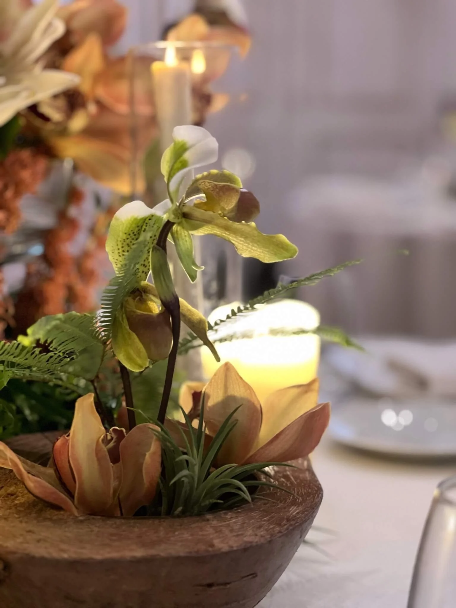 Un centre de table avec une orchidée blanche et verte, des feuilles vertes, des bougies jaunes dans un vase en verre, et un décor de fleurs et de bougies autour.