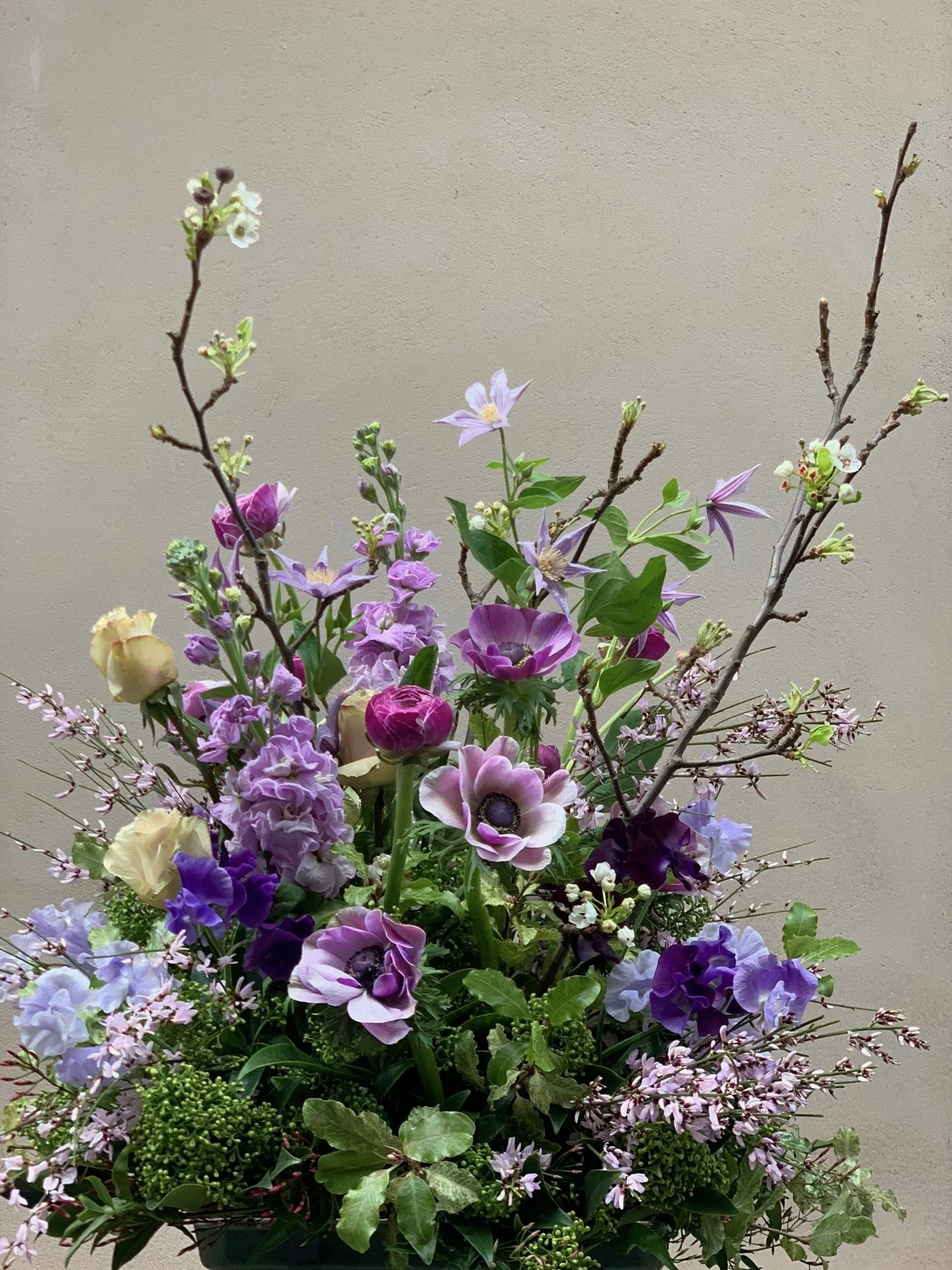 Un bouquet de fleurs colorées avec des nuances de violet, roses et crème, comprenant des branches, des pivoines, des anémones et d'autres floraisons, placé devant un mur beige.