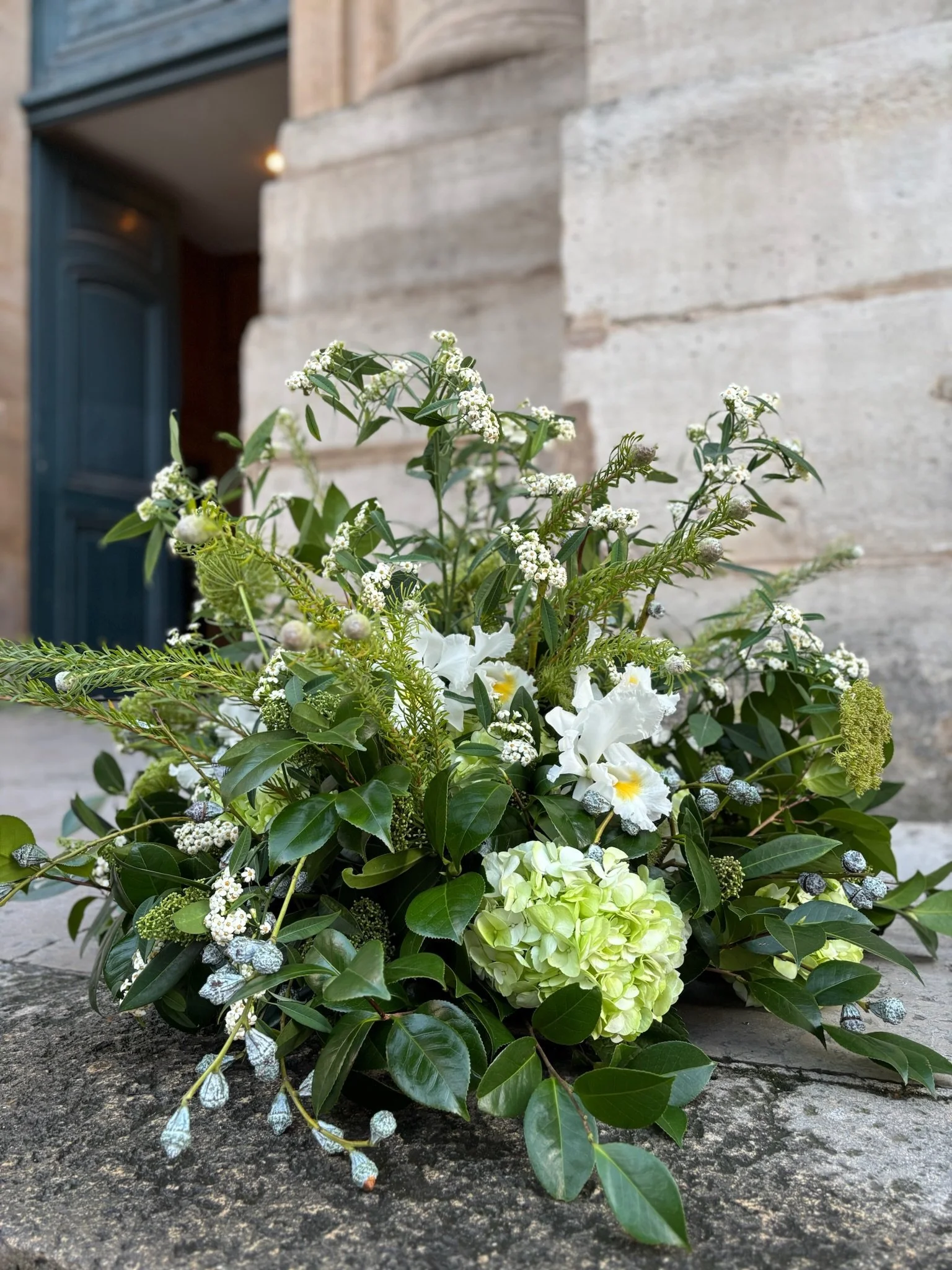Composition florale de deuil avec des fleurs blanches, des feuilles vertes et des baies argentées, posée sur une surface en pierre devant un mur en pierre et une porte bleue.