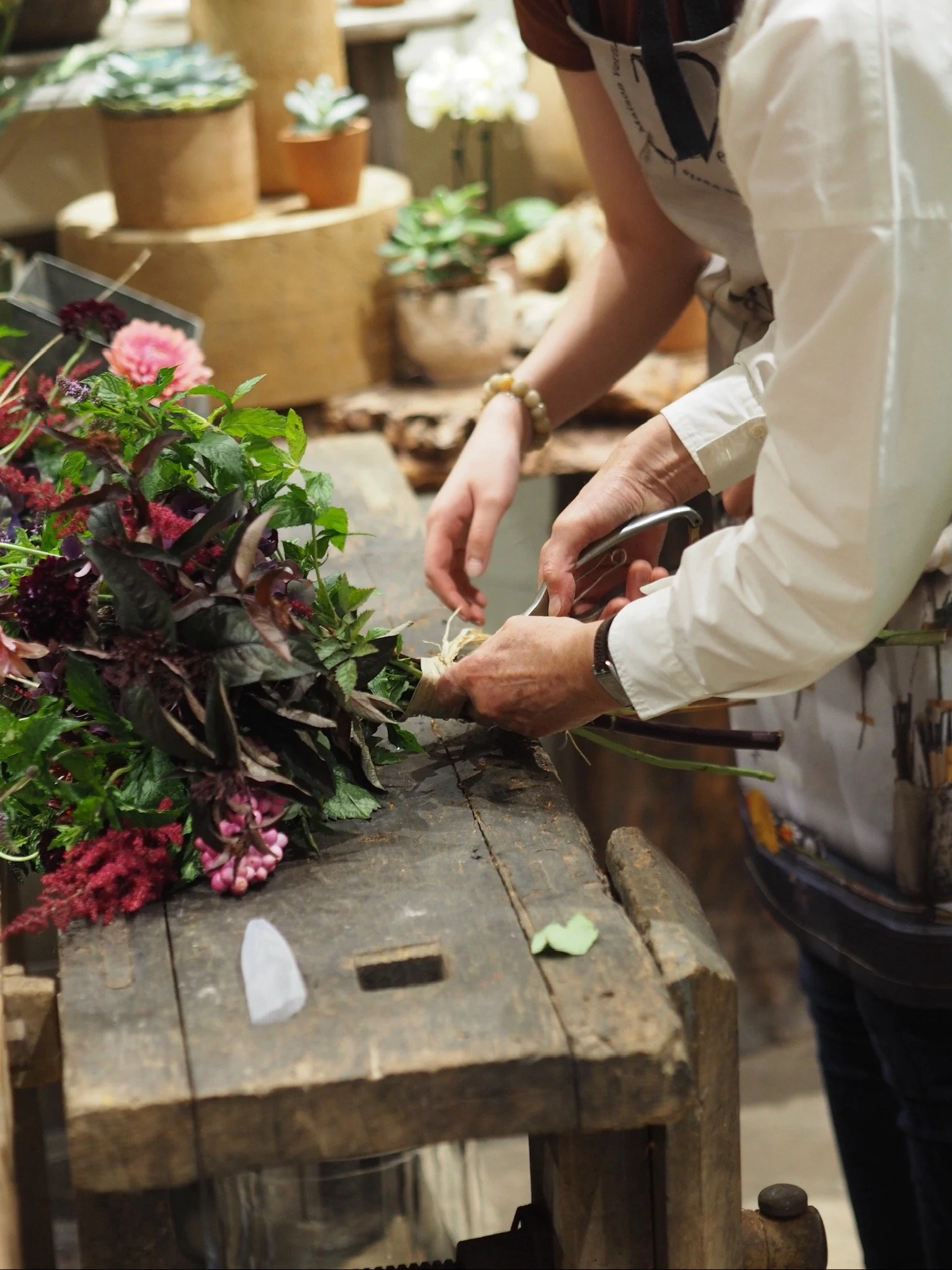 Une personne crée une composition florale avec des fleurs et des plantes diverses sur une table en bois dans une boutique de fleurs.