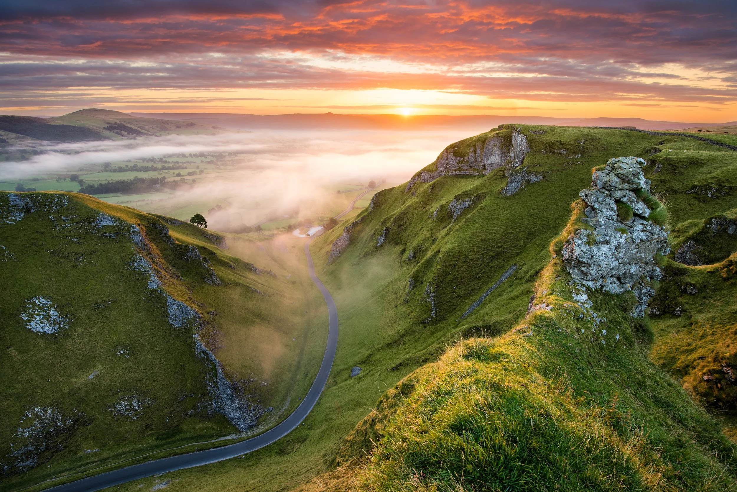 Sunrise over rolling green hills with a winding road, mist in the valley, and a partly cloudy sky.