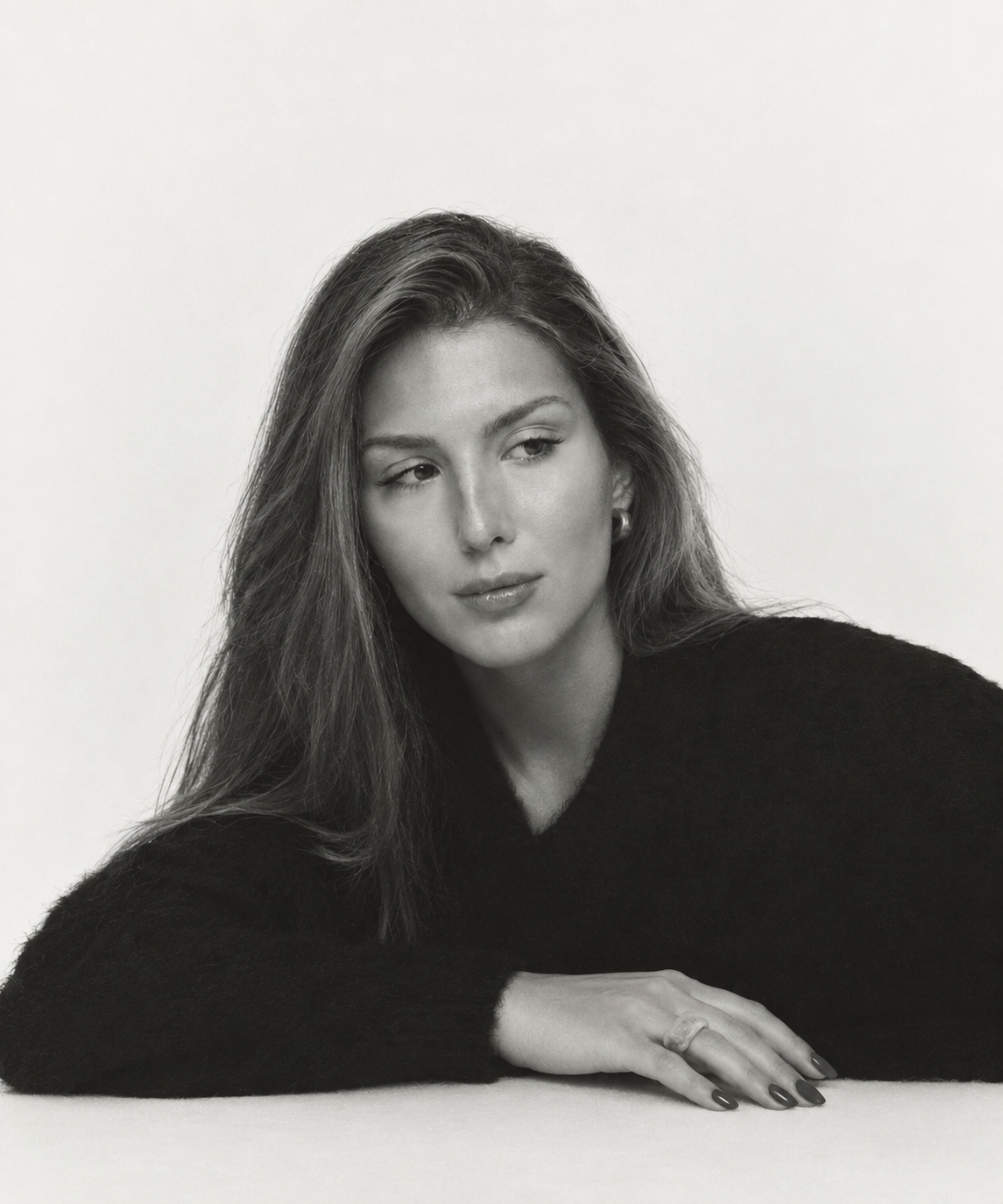 Black and white portrait of a woman with long hair, wearing a dark sweater, resting her chin on her folded arms, with a neutral expression.