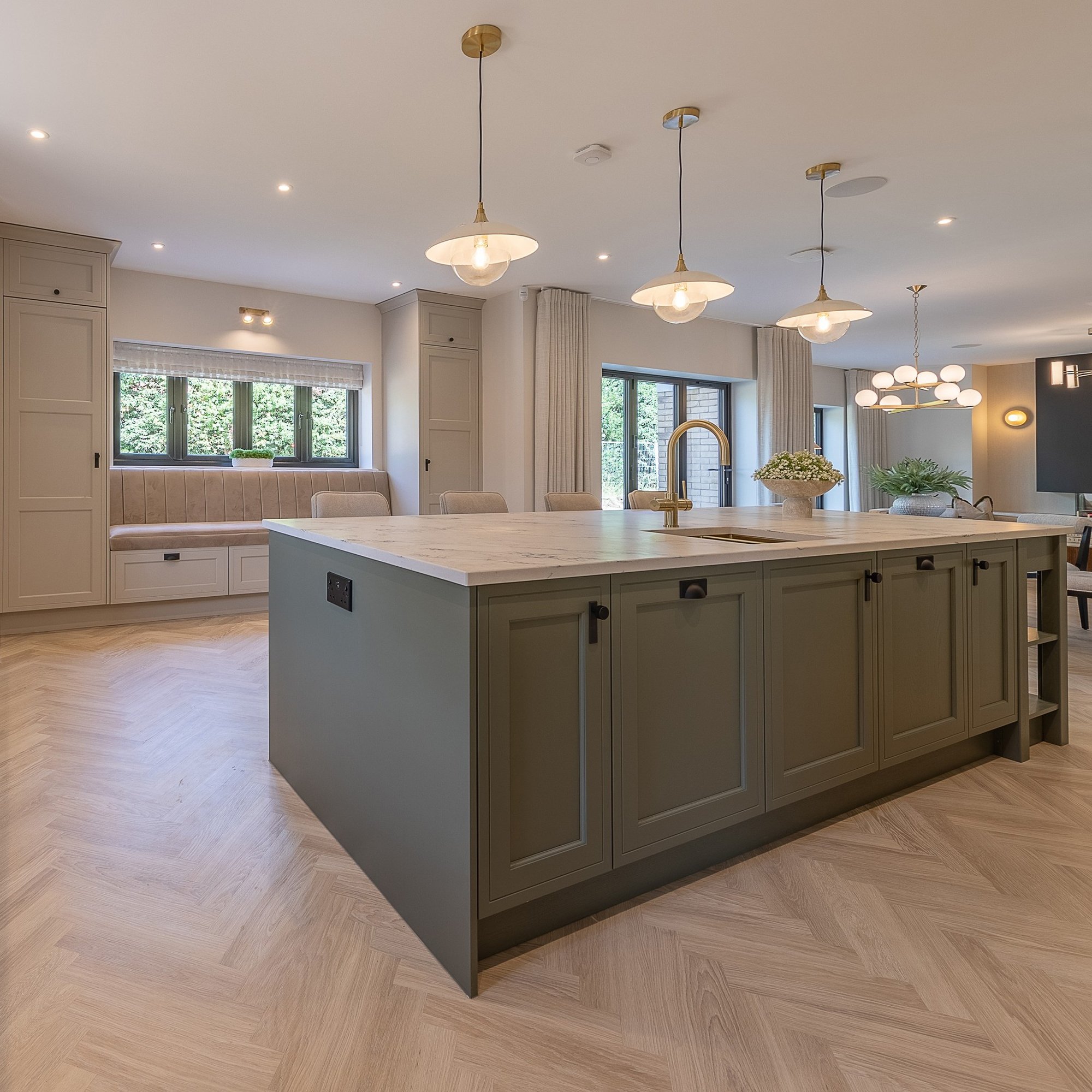 Modern kitchen with an island and gold faucet, featuring pendant lights, large windows, built-in seating, and light wood herringbone flooring.