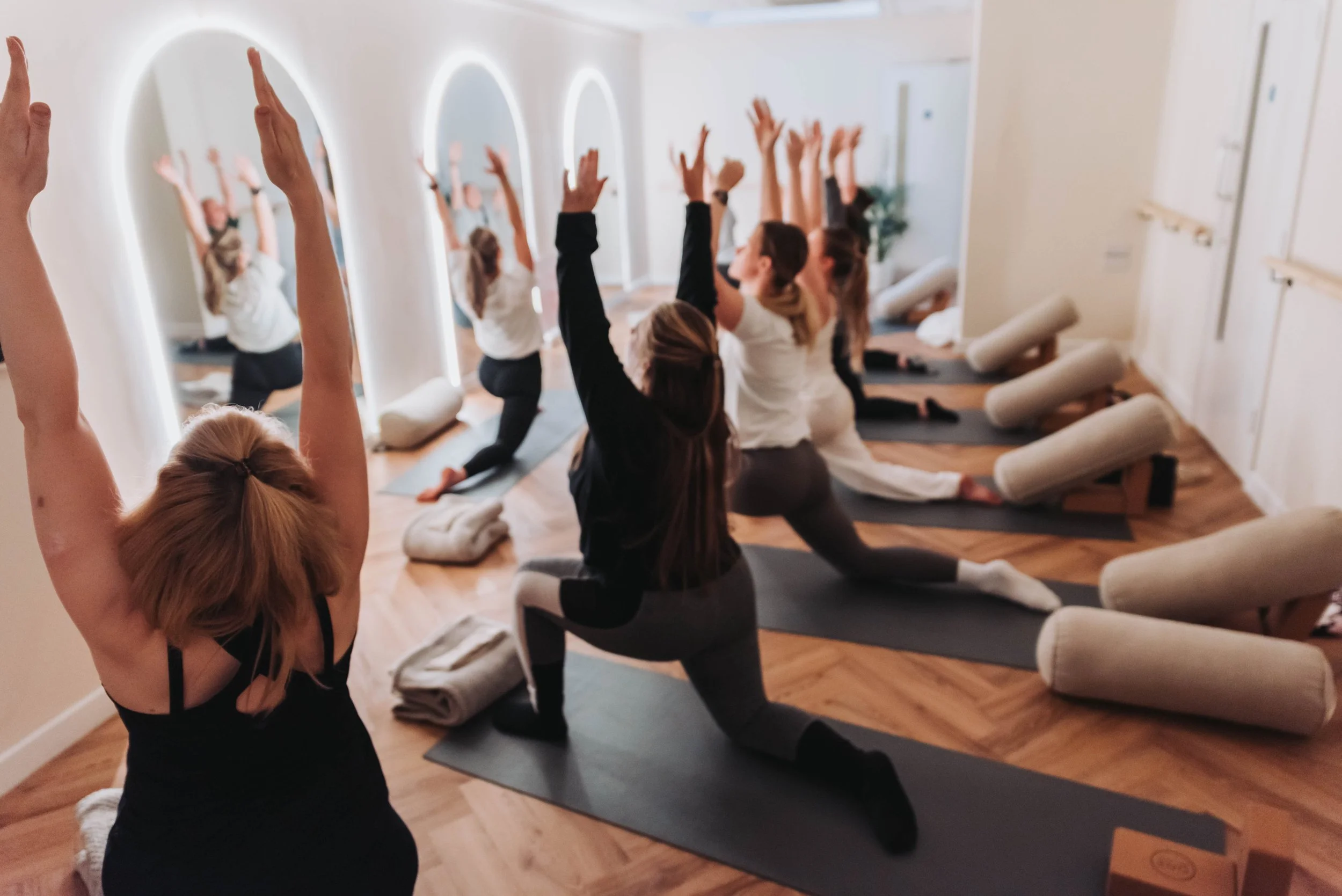 Group of people participating in a yoga class in a well-lit studio with mirrors, practicing a yoga pose with arms raised and kneeling on yoga mats.