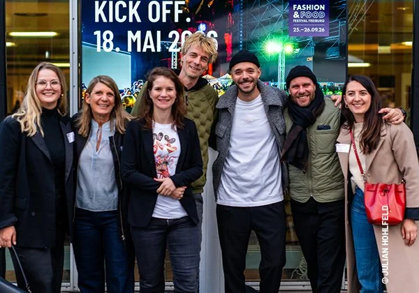 Eine Gruppe von sechs Menschen bei einer Veranstaltung im Freien, einige halten Wein- oder Getränkegläser, im Hintergrund sind weitere Personen und ein modernes Gebäude mit Glasfassade sichtbar.
