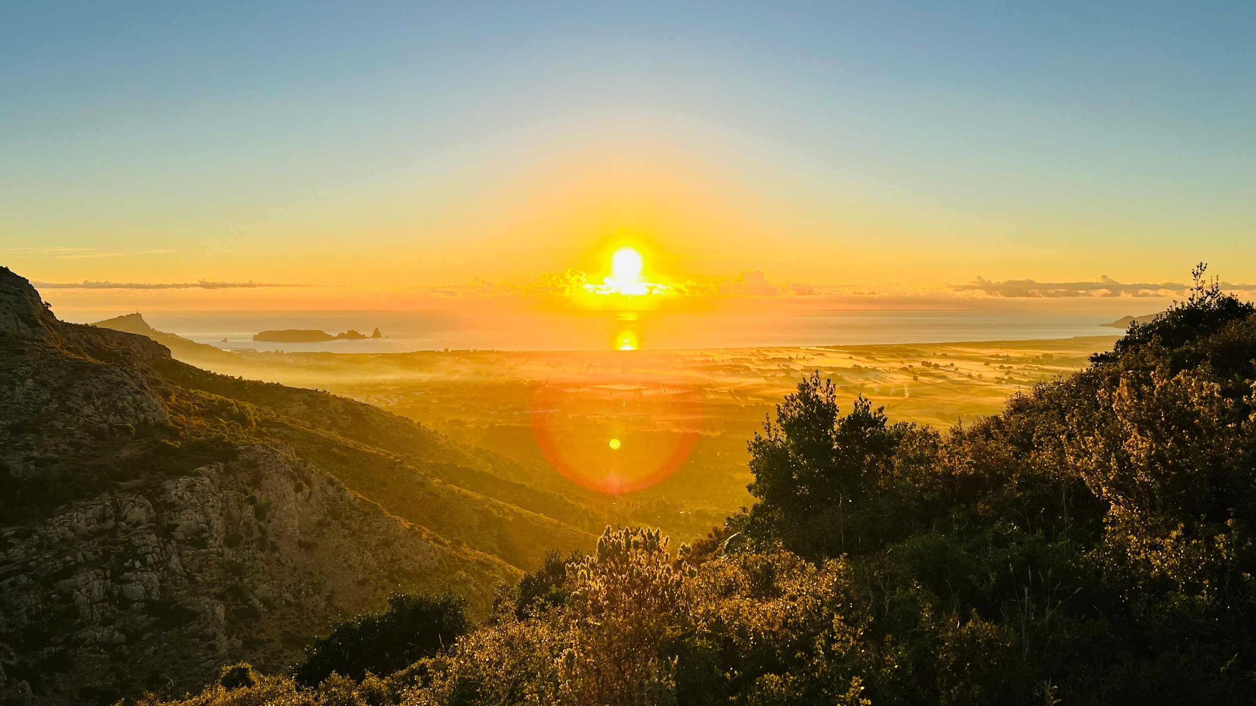 Zonsondergang boven een landschap met bergen, water en groene bomen.