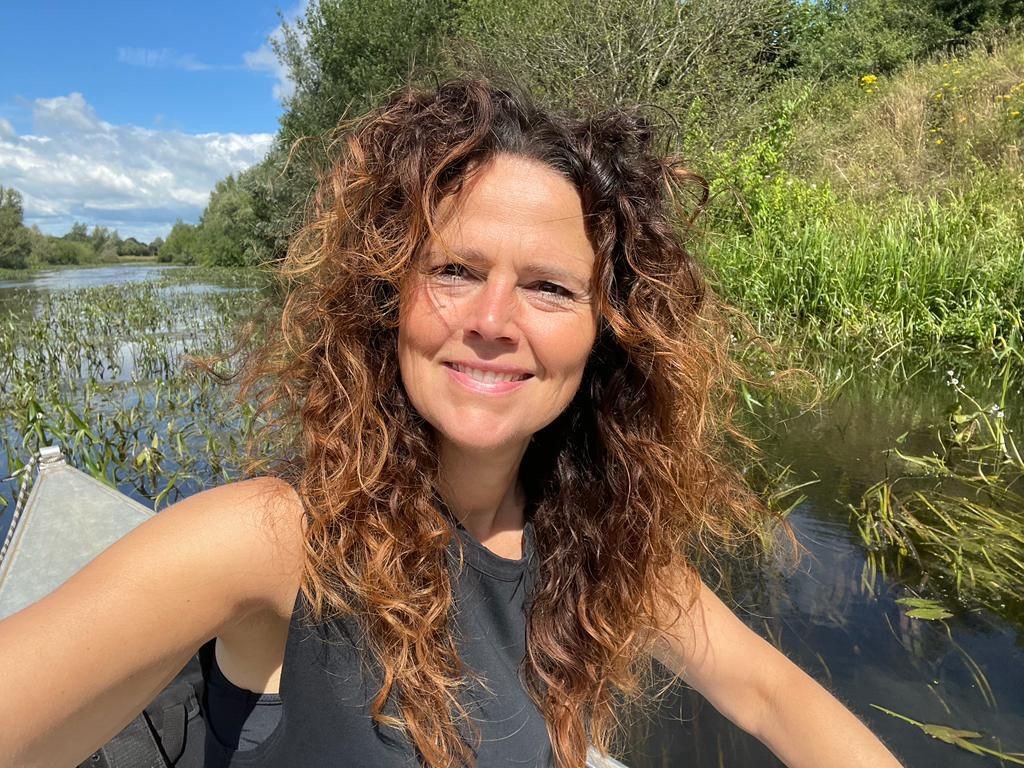 Vrouw maakt selfies in de natuur bij een rivier met groene planten en bomen, blauwe lucht en witte wolken.