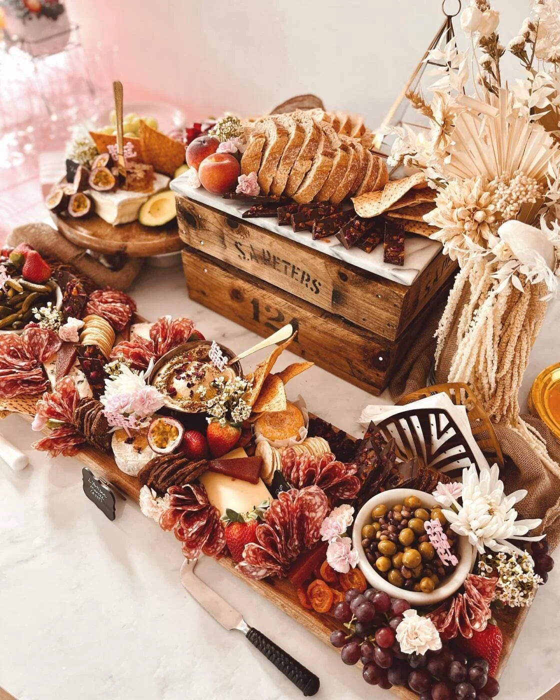 Decoratief kaas- en vleesplateau met fruit en bloemen, plus een broodmand en een andere dessert op de achtergrond.