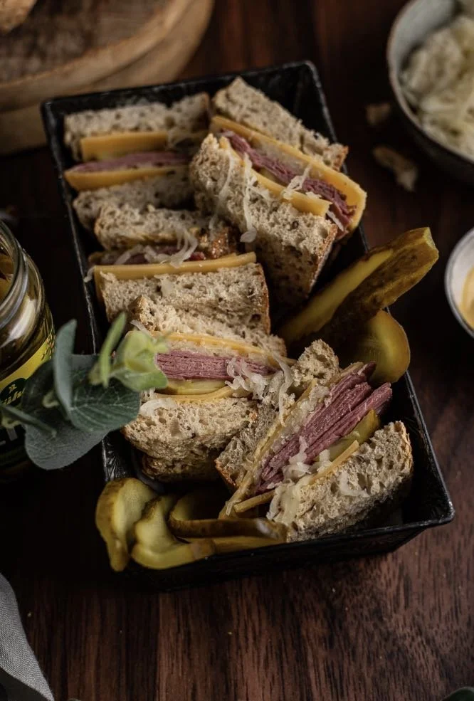 Sandwiches met vlees, kaas en zuurkool in een zwarte doos, met augurken en een drankje ernaast.