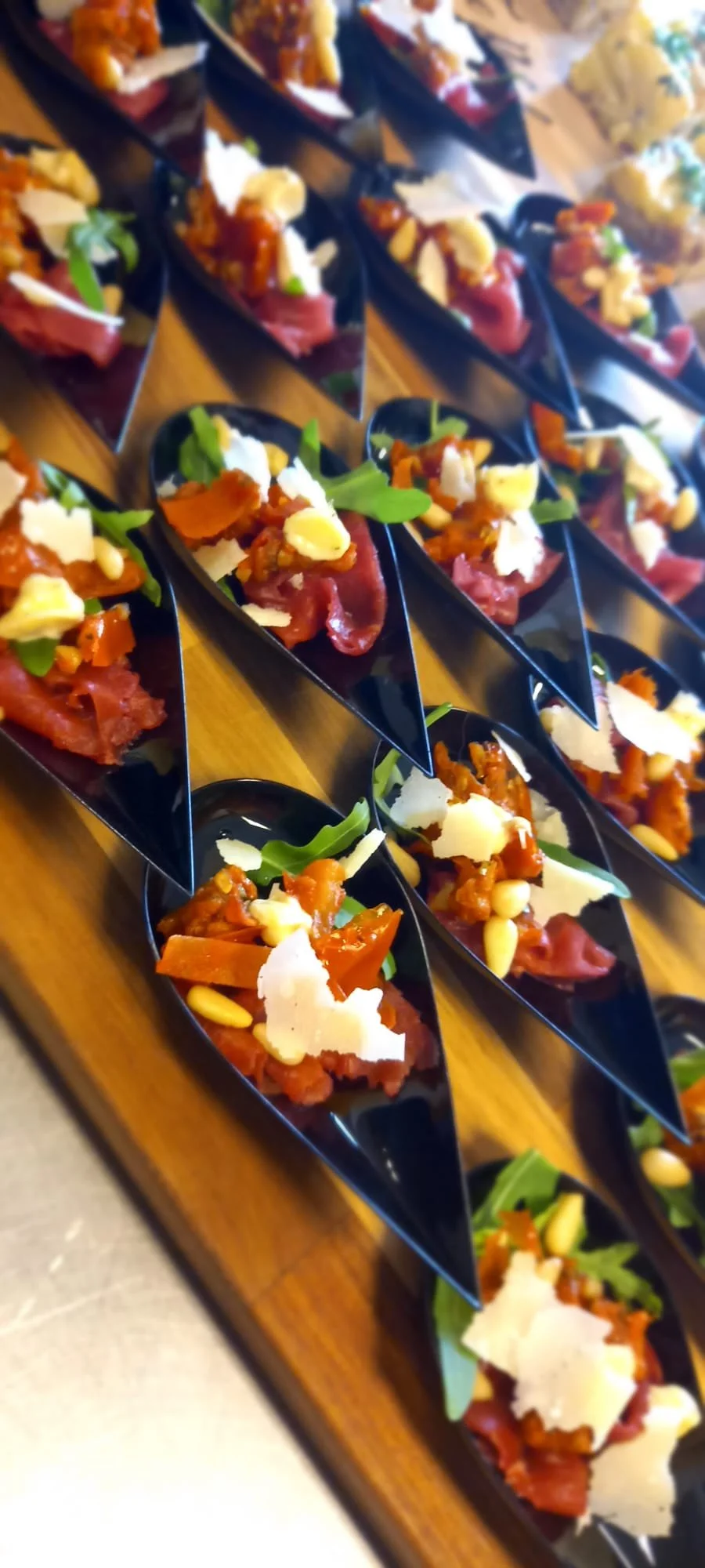 Kleine hapjes met vlees, kaas, groenten en noten op zwarte lepels, uitgestald op een houten dienblad.