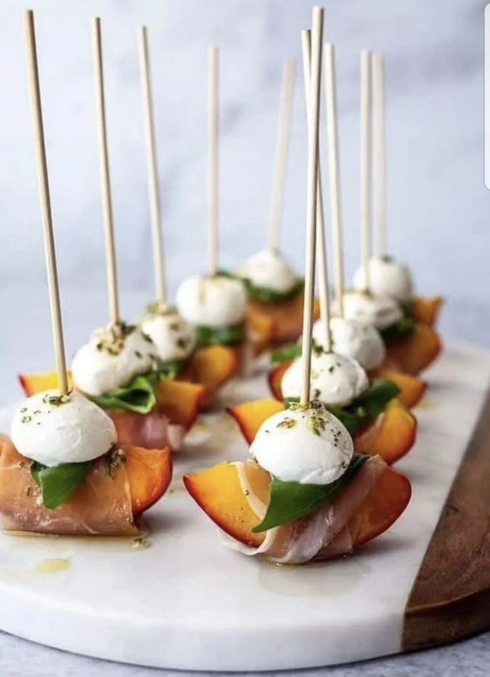 Kleine hapjes met mozzarella, verse kruiden, perziken, en vlees op een houten en marmeren serveerschaal.