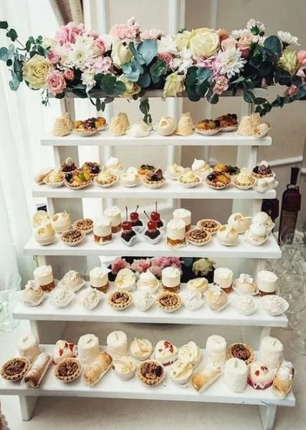 Een wit fleurig bloemenarrangement boven een wit koekjesrek met diverse kleine zoete gebakjes en desserts.