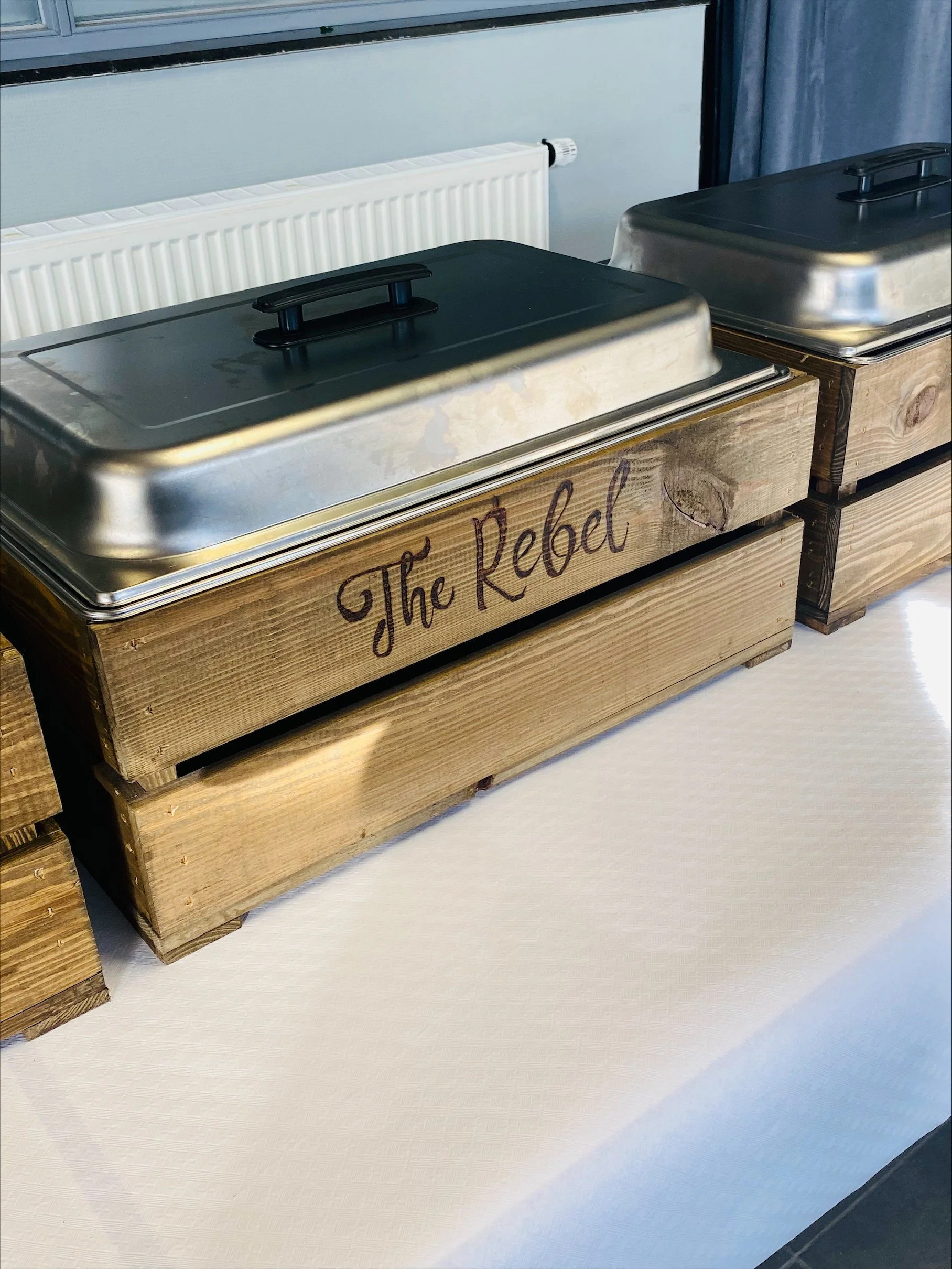 Twee houten kisten met metalen deksels en een inscriptie 'The Rebel' op de voorkant, geplaatst op een witte tafel.