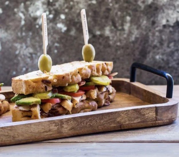 Twee sandwiches met kip, komkommer, tomaat en olijven op een houten dienblad.