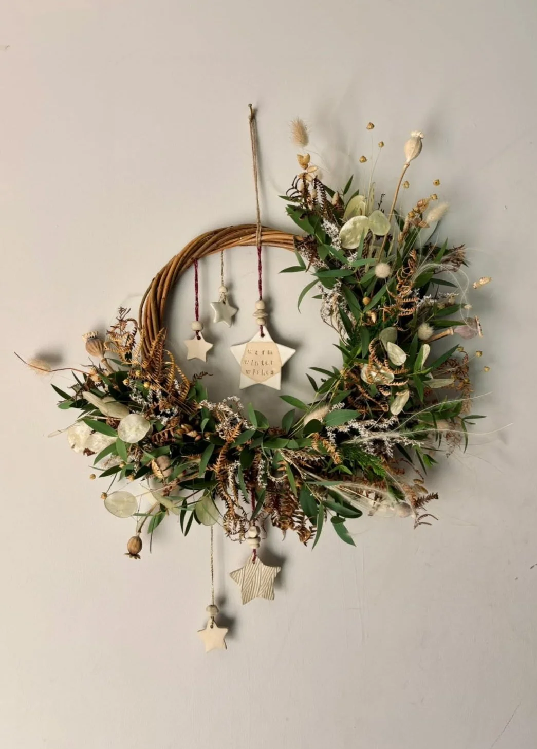 A decorative wreath made of intertwined branches, adorned with dried green leaves, beige flowers, and small white fluffy accents, hanging on a plain white wall. The wreath is decorated with hanging star ornaments, some with text, and small beads.