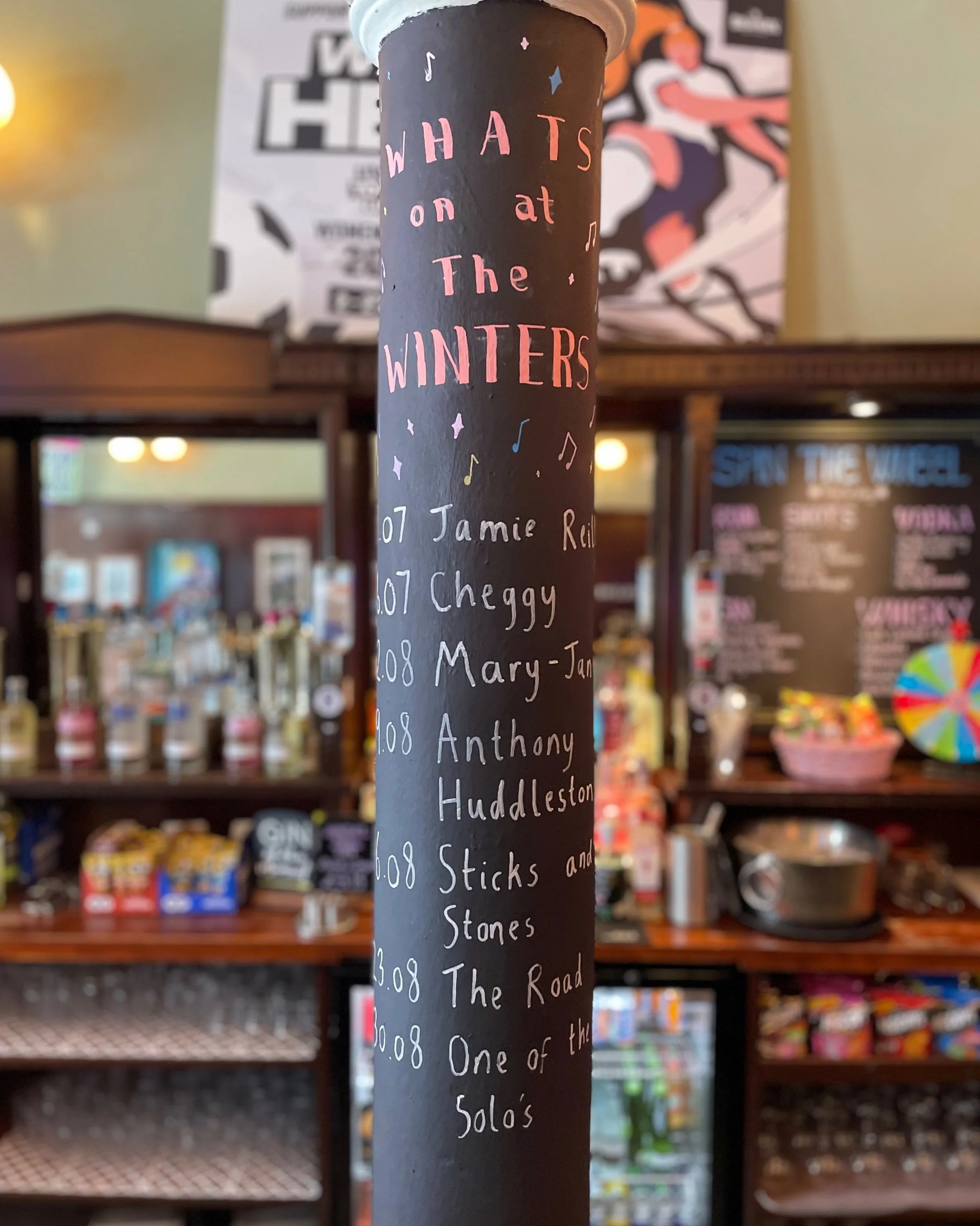 A black vertical pole with pink and white handwritten text listing songs and performers, with a background of a snack bar and a large colorful wheel.