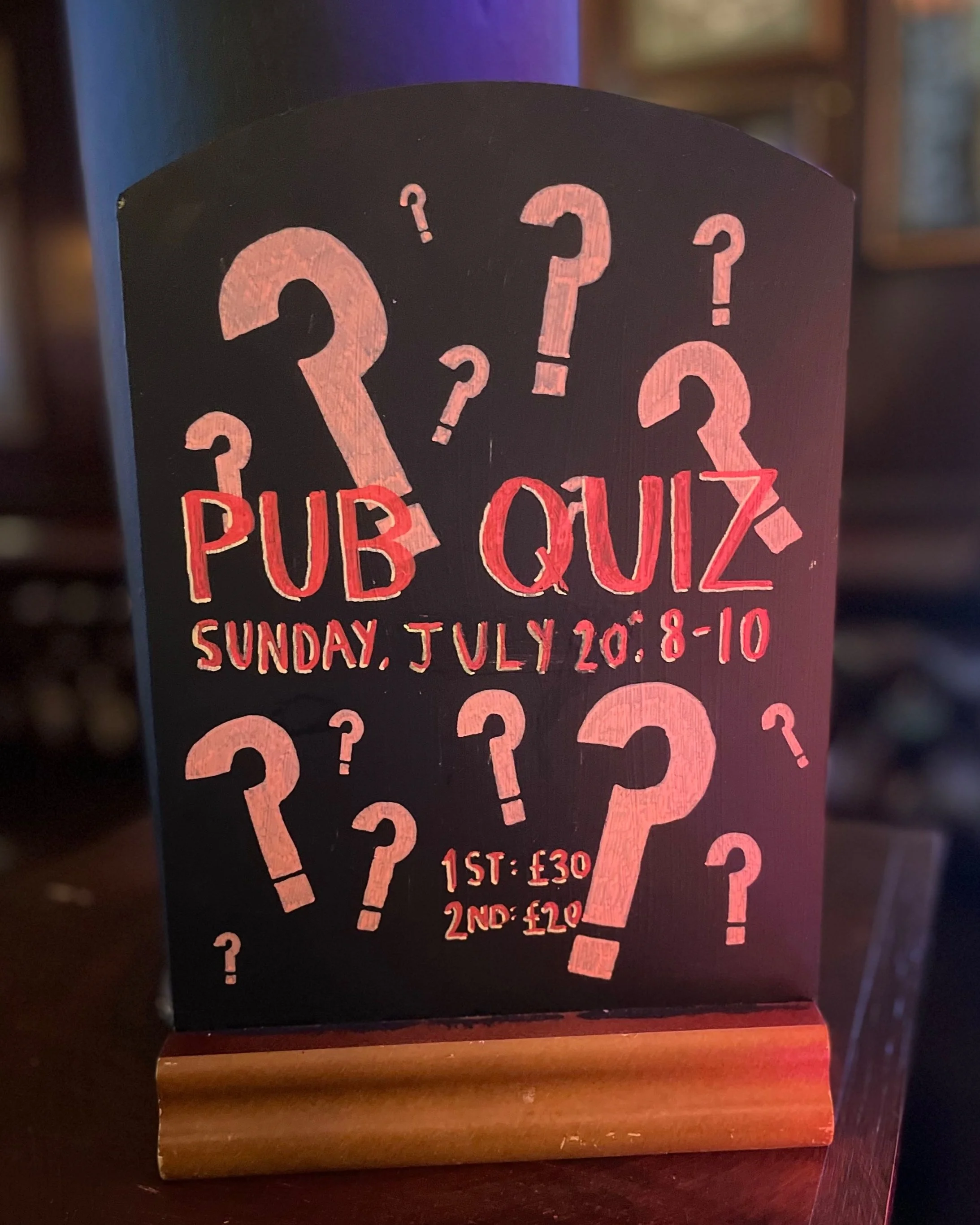 Blackboard sign advertising a pub quiz on Sunday, July 20 from 8 to 10 pm with two quiz entry prices: £30 for the first entry and £20 for the second. The sign features various pink question marks.