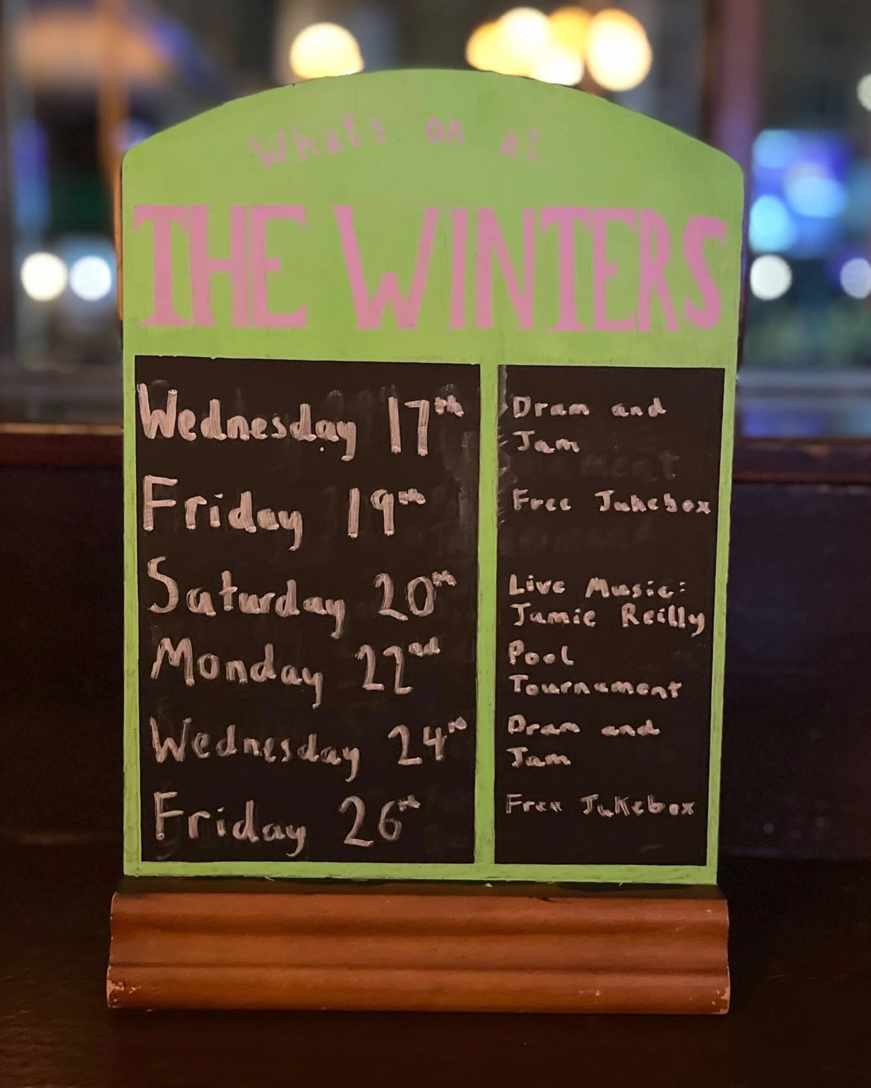 A green standing signboard with the header 'What's on at THE WINTERS' in pink letters, listing events scheduled from Wednesday the 17th to Friday the 26th, including live music by Jamie Reilly, a pool tournament, and free takeout by Dear and Jim.