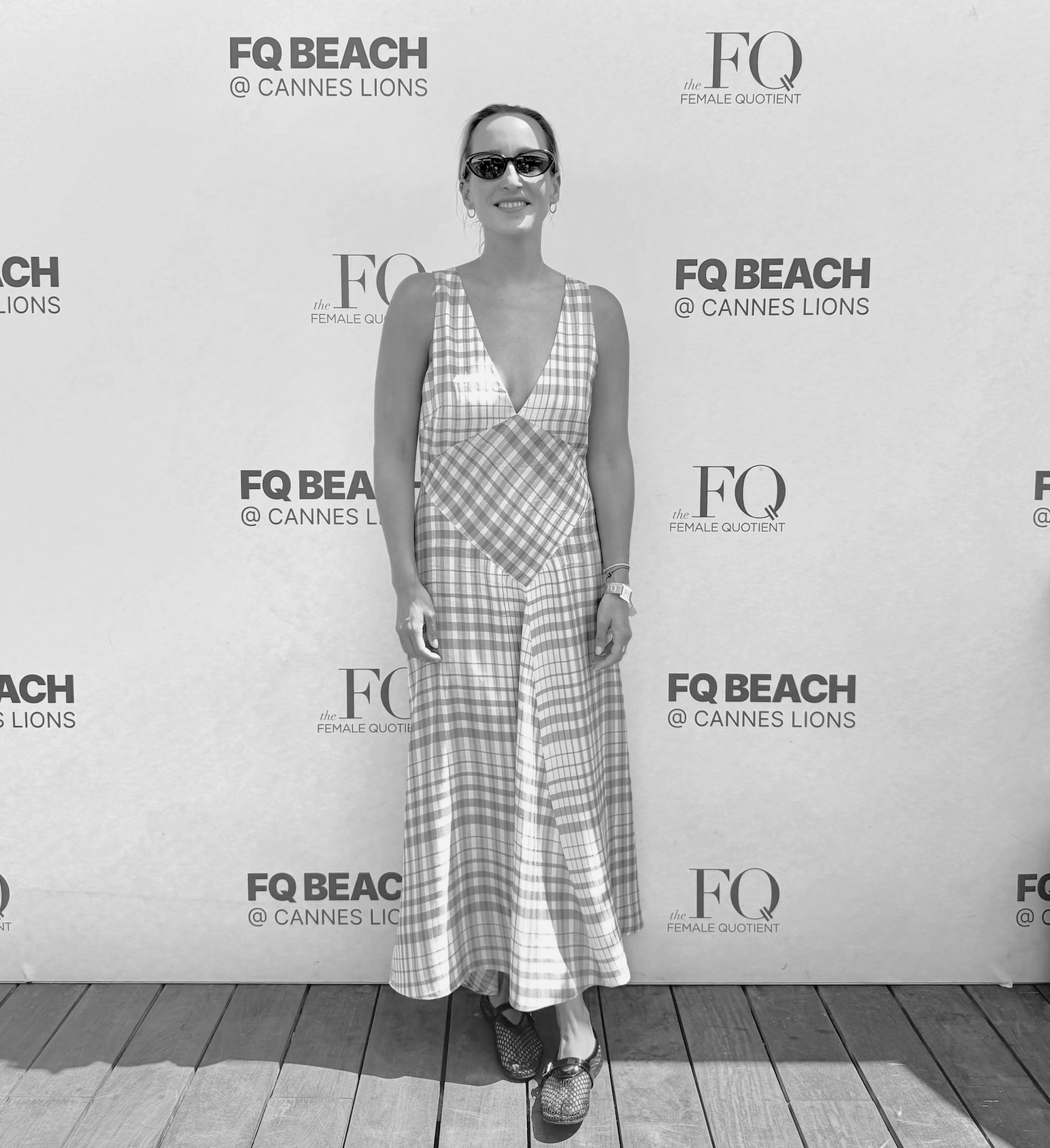 A woman standing on a wooden floor in front of a white backdrop with the logos of FQ Beach and Female Quotient, dressed in a checkered sleeveless top and wide-legged patterned pants, wearing sunglasses and smiling.