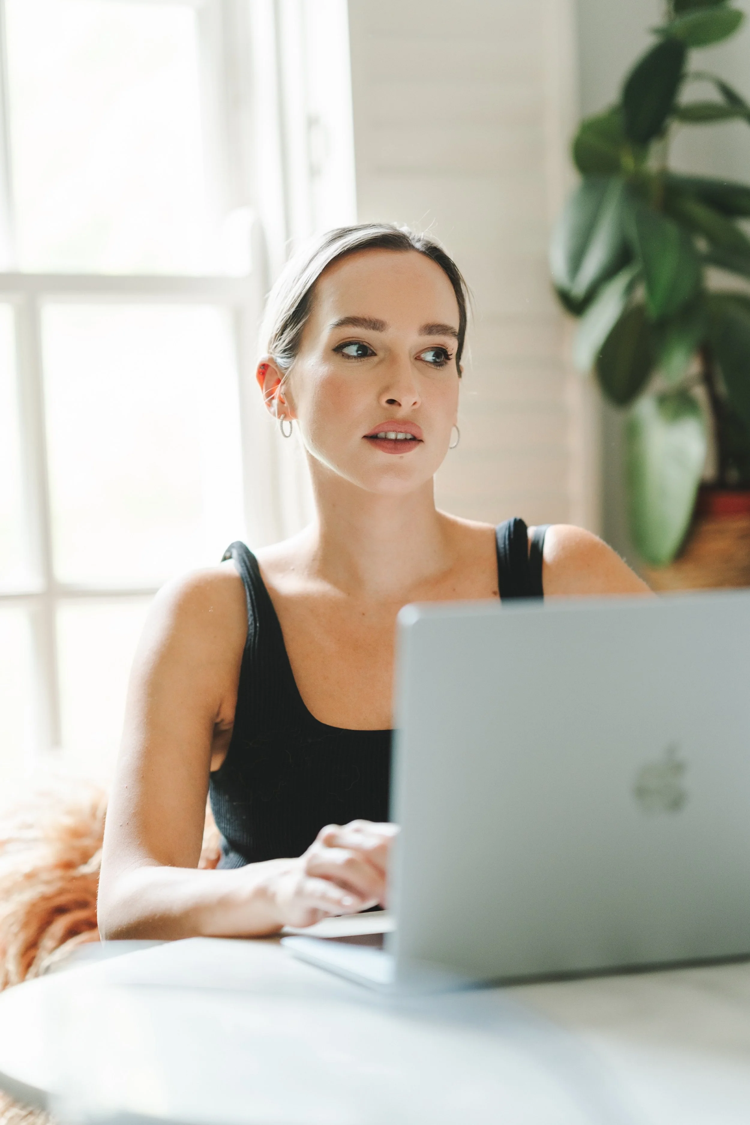 A woman with short brown hair and hoop earrings sitting at a desk and looking at a laptop screen, with natural light coming through a window behind her and a large green plant nearby.