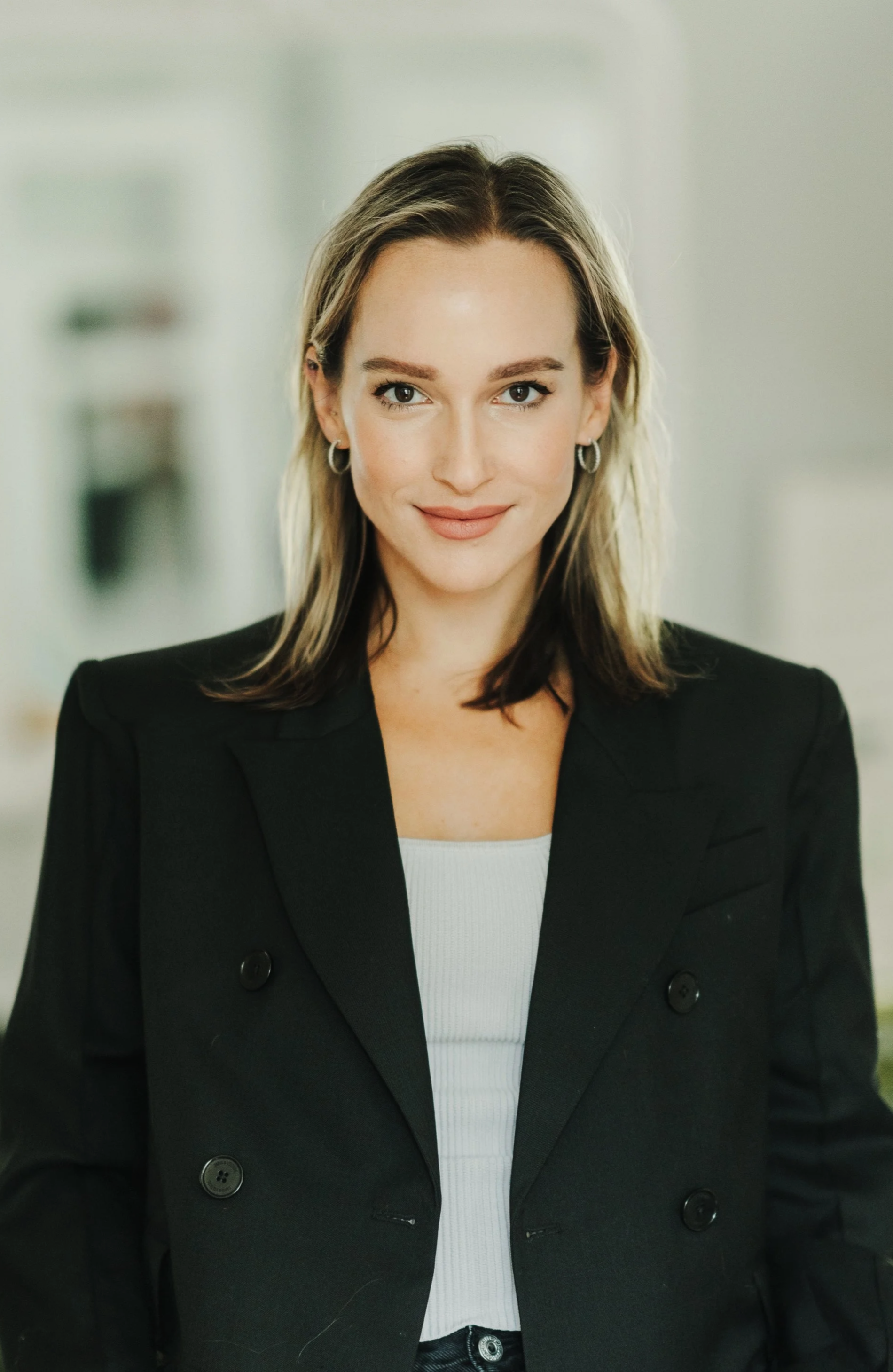 A woman with shoulder-length light brown hair, wearing hoop earrings, a white top, and a black blazer, standing in a professional indoor setting.