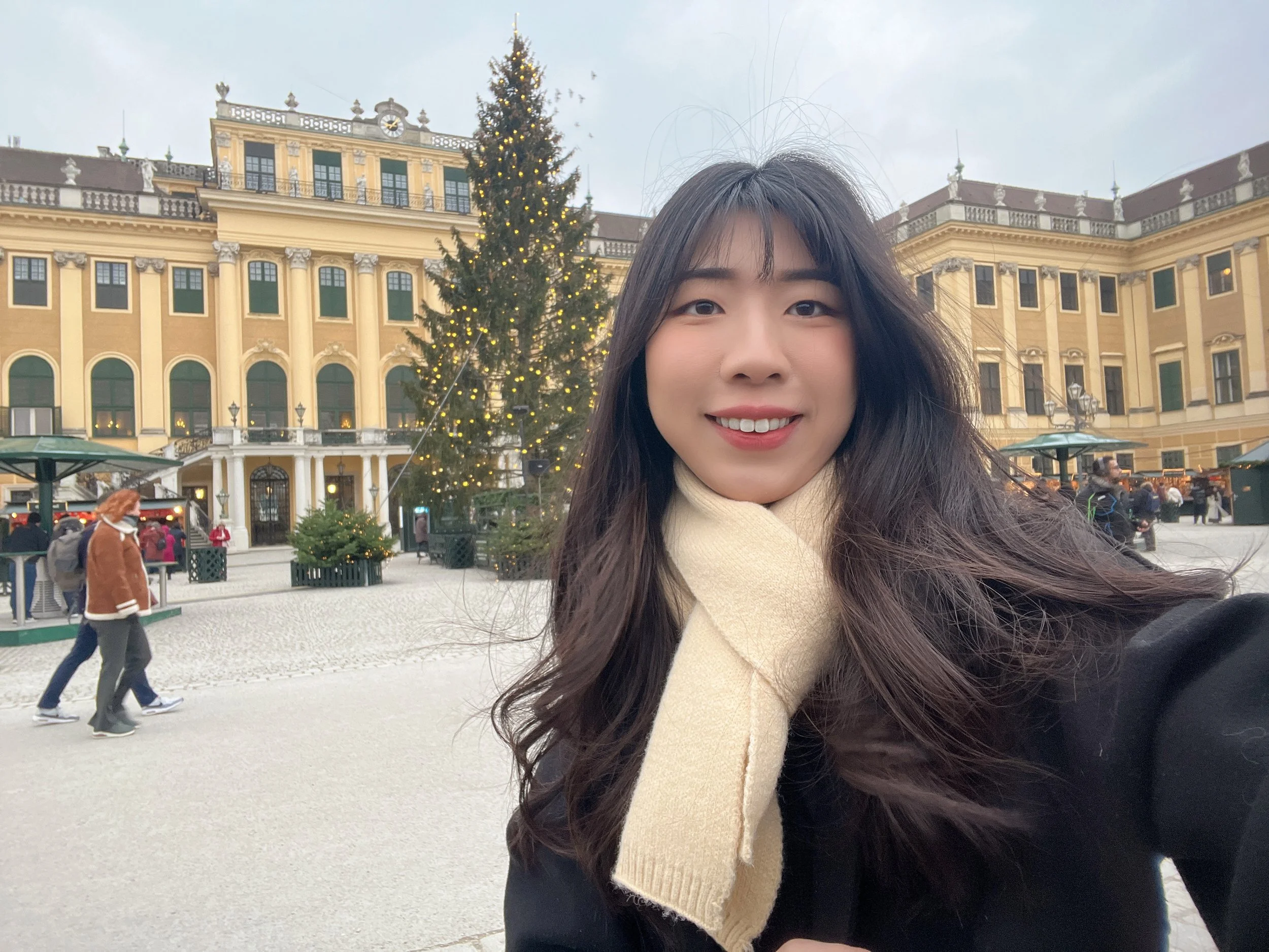 Hazel Hsinchi Hsu looks at the camera with a christmas tree behind her, she has long brown hair, a cream scarf and black jacket